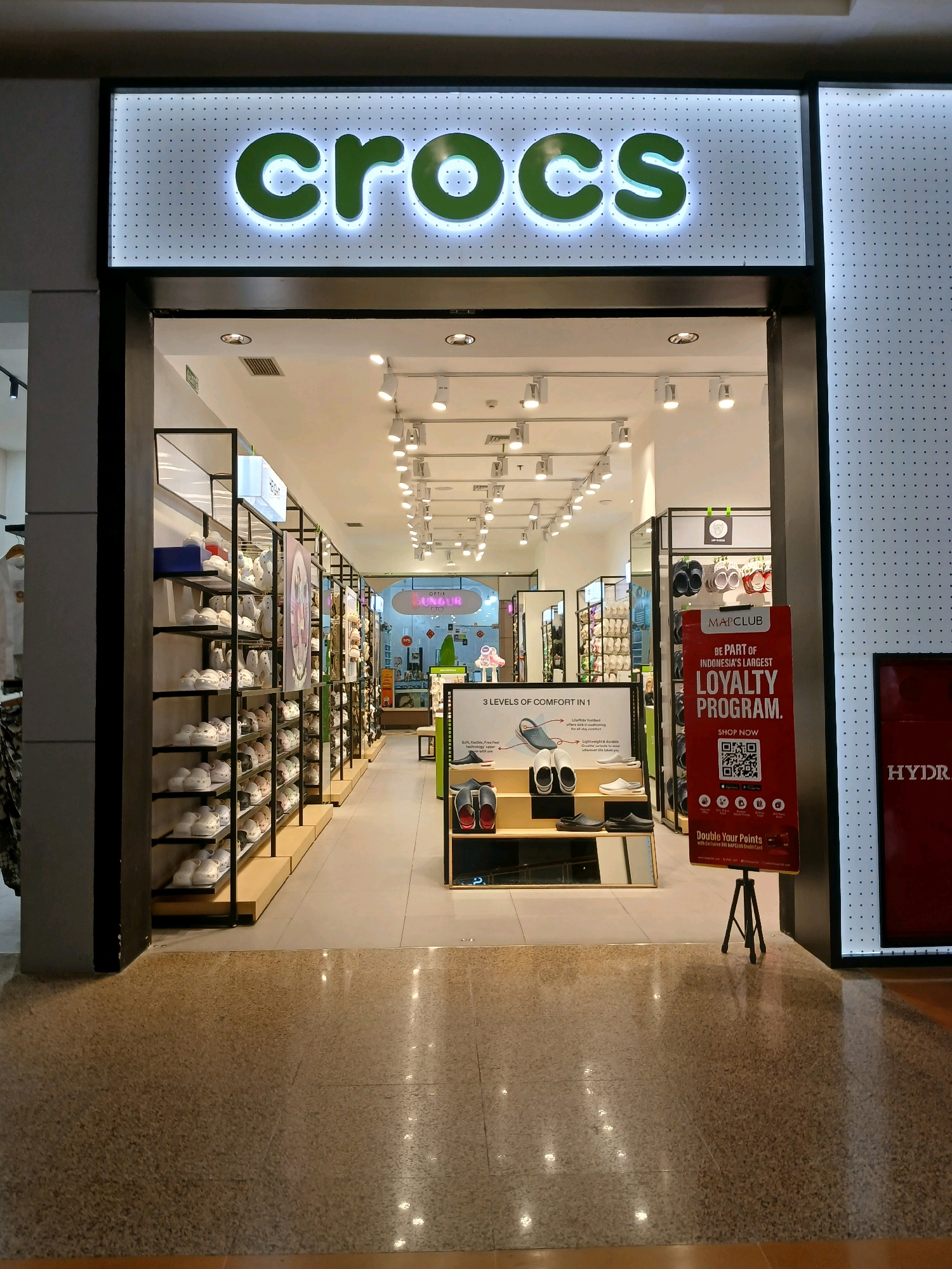 The image shows the entrance to a Crocs store located in North Jakarta, Indonesia. The store is well-lit and displays a variety of Crocs footwear on shelves. A large, illuminated sign above the entrance reads "crocs" in green lettering. To the right of the entrance, a red promotional stand for "MAPCLUB" and its loyalty program is displayed. The floor is made of speckled tiles, and the overall environment appears to be a shopping mall. There are no people visible in the image.