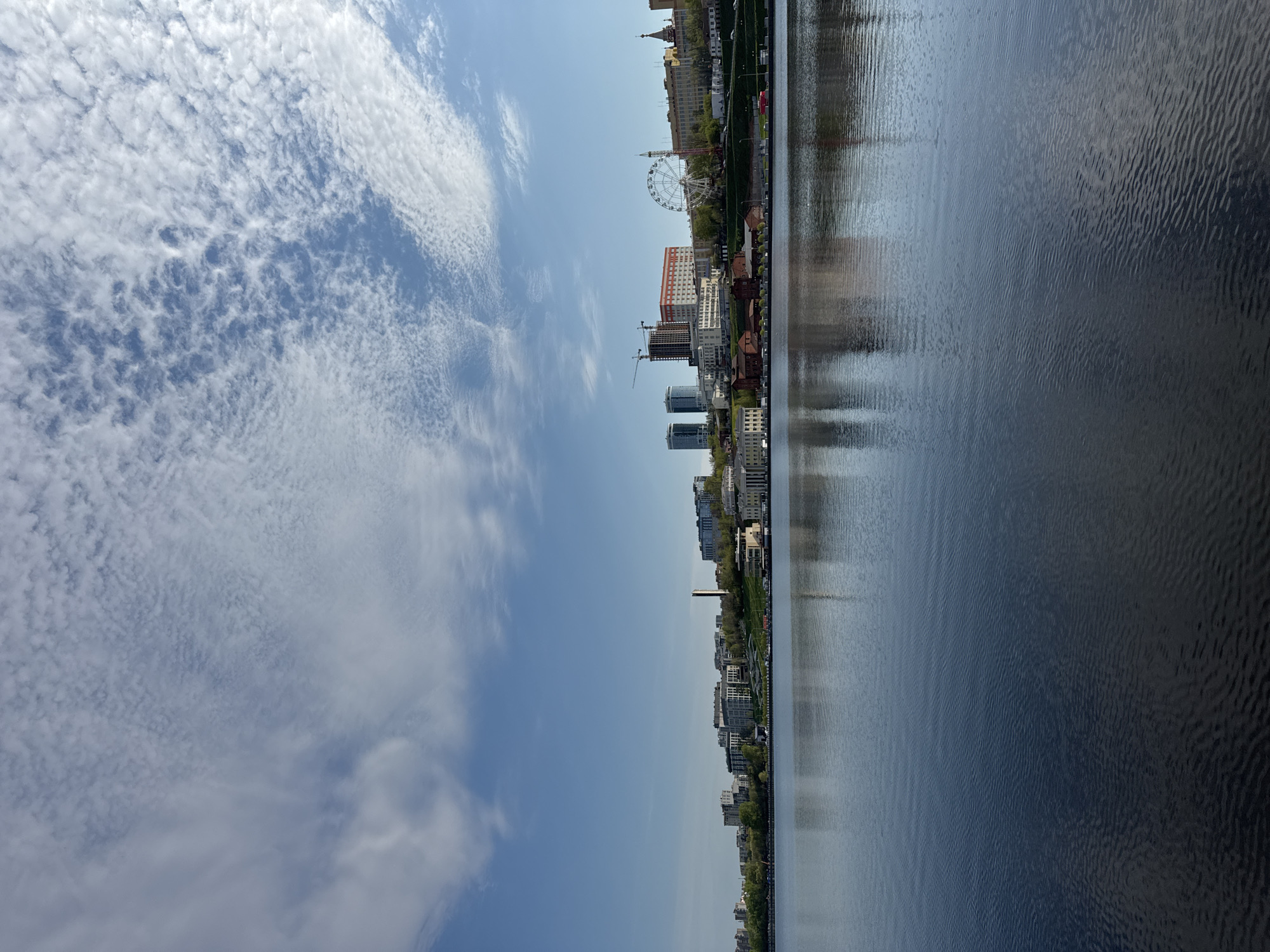 The image depicts a view in Izhevsk, Russia, featuring the city skyline reflected in a body of water. The sky is partly cloudy. A Ferris wheel is visible amongst the buildings, indicating an amusement park or recreational area along the waterfront. Construction is underway, with a crane present at a construction site. The water's surface displays ripples and reflections of the city's architecture and sky. Vegetation lines the waterfront.