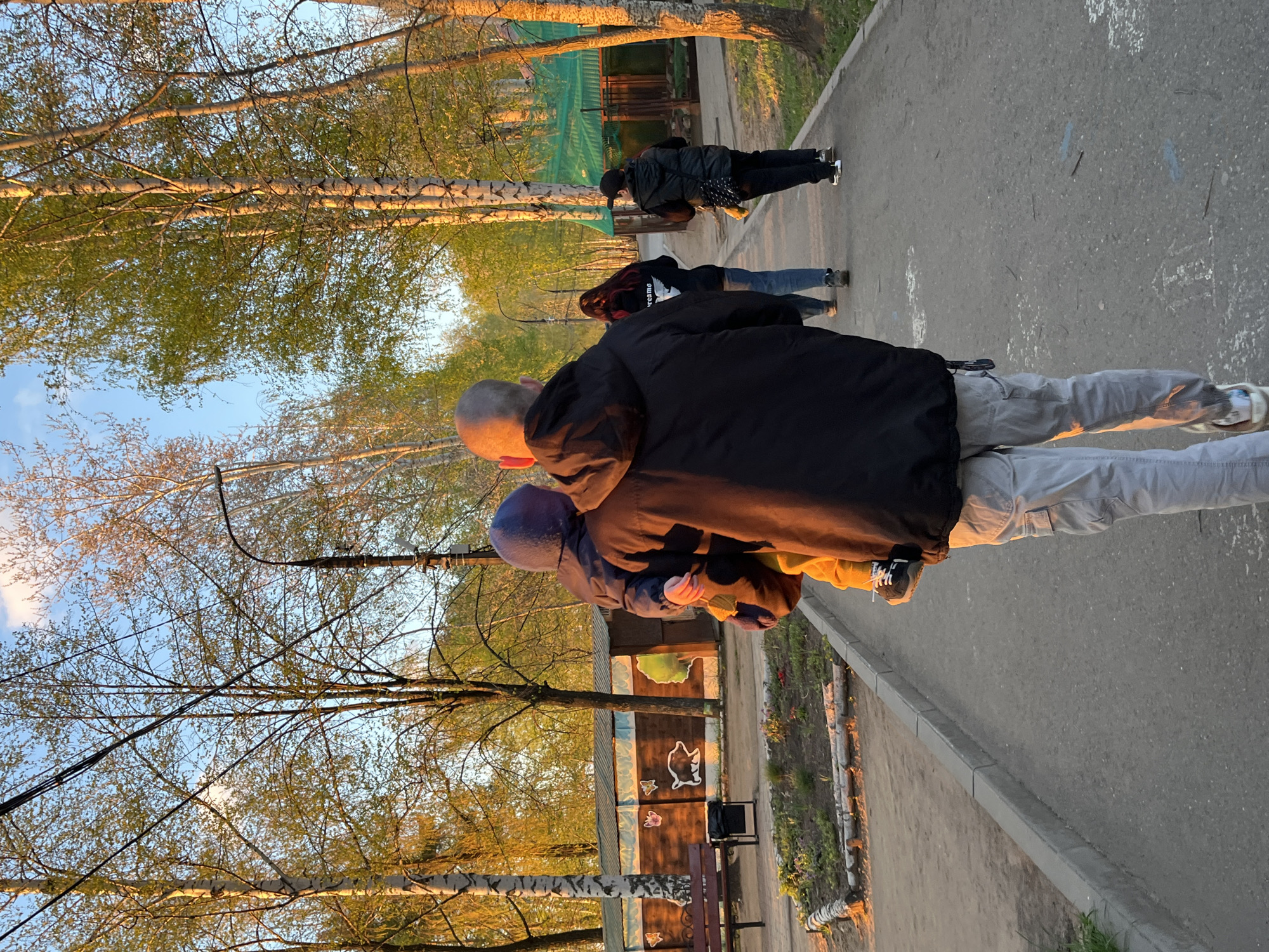 A father is carrying a child on his back while walking on a paved path in a park in Chaikovsky, Russia. The father is wearing a black jacket and grey pants. The child is wearing a blue hooded jacket and is nestled against the father's back. Other people are walking ahead of them on the path. Trees with new spring leaves line the path, and sunlight filters through the branches. A fence with cartoonish animal drawings on it is visible in the background. A bench and trash can are positioned next to the fence. The scene appears to be captured during the golden hour, as the lighting is warm and casting long shadows.