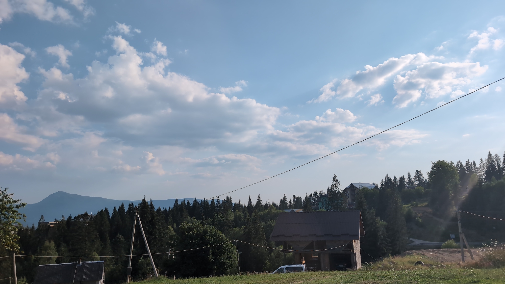 The image shows a landscape with a clear blue sky filled with white, puffy clouds. In the foreground, there is a wooden structure with a dark, sloped roof. To the left of this structure, another smaller building with a metal roof is partially visible. A white van is parked near the wooden structure. A power line stretches diagonally across the upper portion of the image. Behind the buildings, a dense forest of pine trees covers the hillside, with a single, larger mountain visible in the distant background. The location is identified as Yablunytsya, Ukraine.