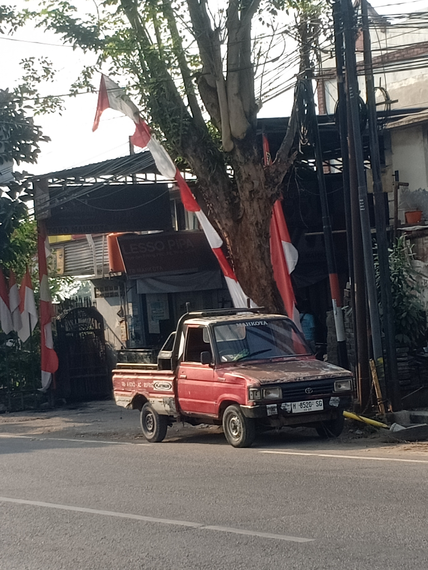 An overcast, daytime scene depicts a red, weathered pickup truck parked on the side of a paved road in Semarang, Indonesia. The truck, with its license plate reading "H-8520-SG", is positioned in front of a low-slung building with a corrugated metal roof. The building has a sign that reads "ESSO PIPA" in large white letters and "3D MAHKOTA" underneath. Indonesian flags, red and white, are visible on poles near the building and draped from a tree. The tree's trunk is thick and gnarled, with branches extending overhead. A tangle of power lines runs overhead and is supported by utility poles. To the right of the building, a narrow alleyway is visible, and a small orange bucket sits on the ground. The road in front of the truck has a white line marking its edge. The scene appears to be a commercial or roadside establishment.