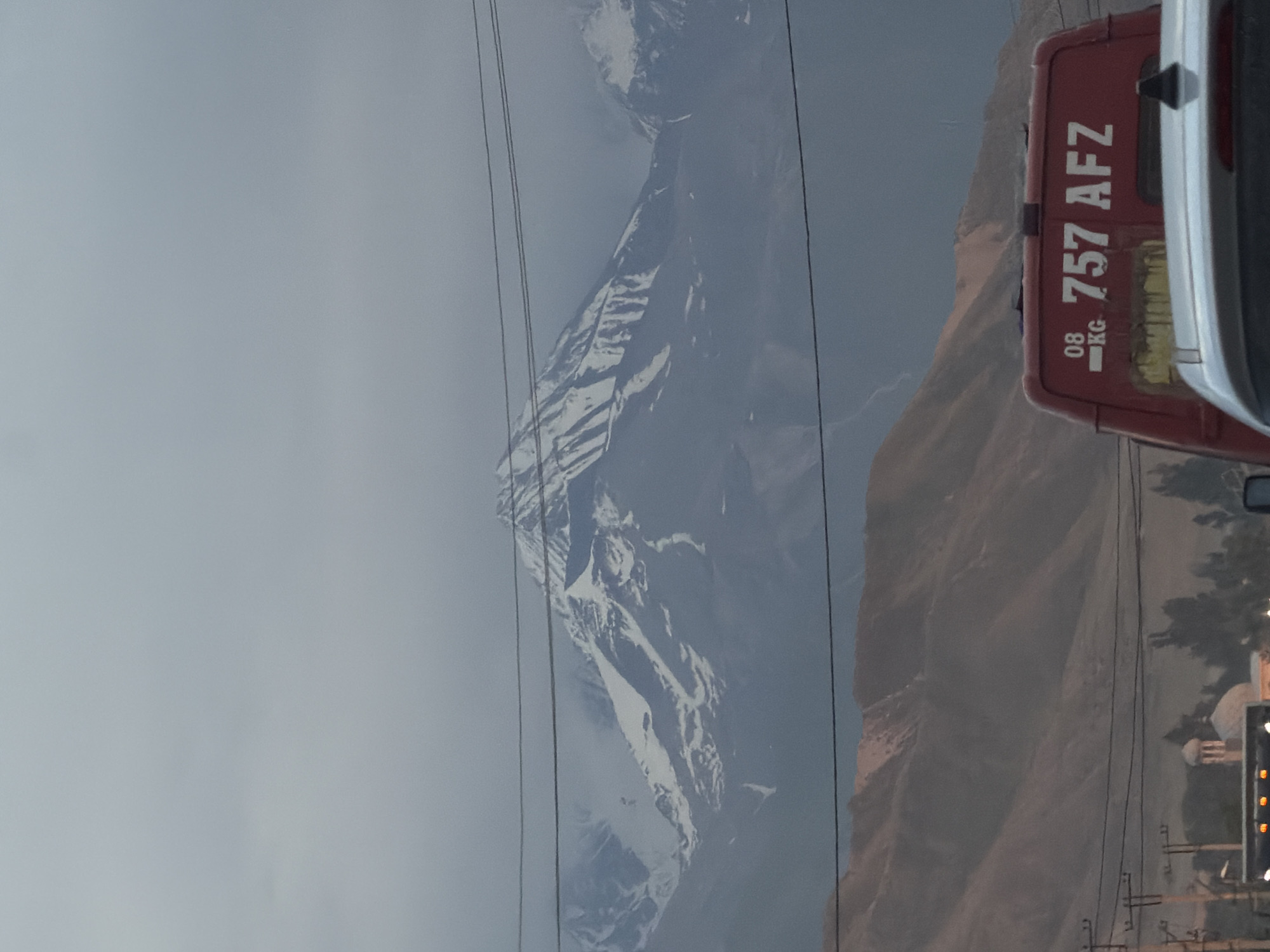 A wide shot captures a majestic, snow-capped mountain range under a cloudy sky. In the foreground, power lines stretch across the frame, obscuring the view of the mountains. Below the mountains, a range of brown hills is visible. A red van with white lettering, "757 AFZ", is parked in front of a white car. The van has "08 KG" printed on its rear. In the distance, a domed building, possibly a mosque, is nestled among trees. The overall scene suggests a rural or semi-urban setting at a distance from a major city.