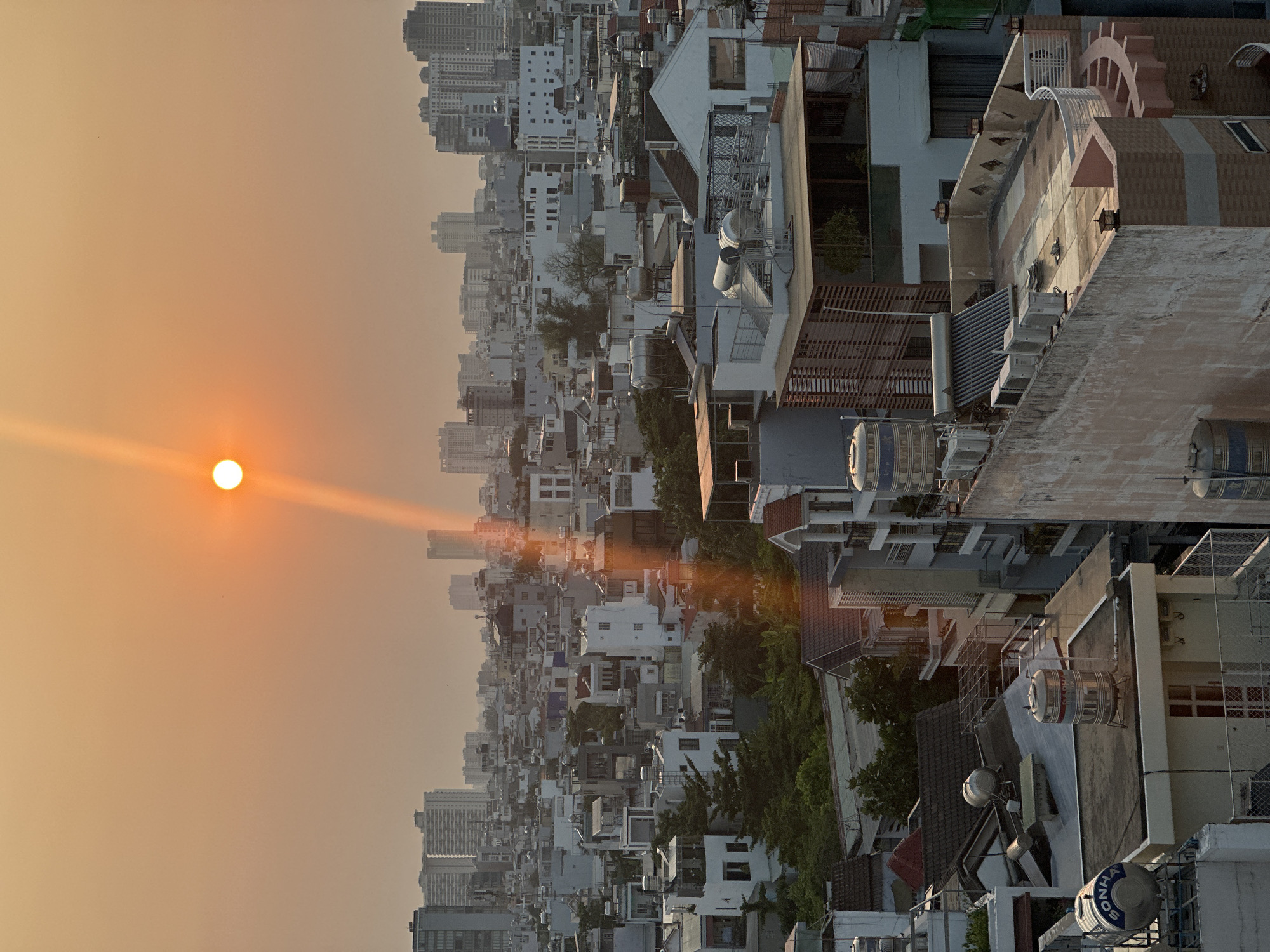 The image shows a cityscape in Ho Chi Minh City, Vietnam, at sunset. The sun is a bright, orange sphere in the sky, with a defined light ray extending towards the urban landscape. The urban area is comprised of a dense cluster of buildings, ranging from low-rise residential structures to taller skyscrapers in the background. The buildings are primarily white or light-colored, creating a visually uniform appearance. A variety of rooftops and architectural details are visible, including water tanks, terraces, and rooftop structures. No people are visible. The environment is characterized by hazy, atmospheric conditions that obscure distant details and give the scene a warm, golden hue.