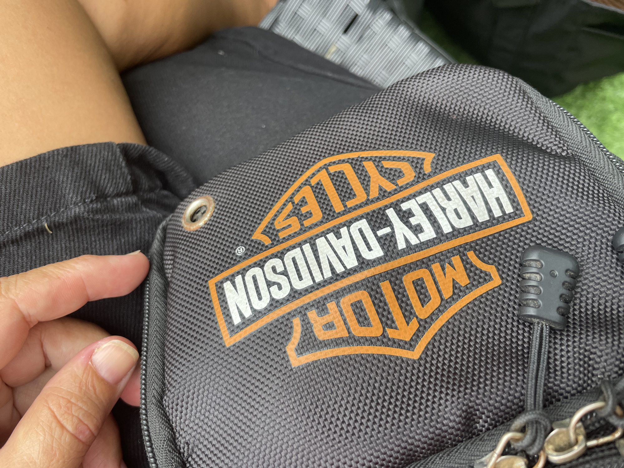 A close-up shot shows a person's hand near a gray Harley-Davidson bag. The bag features the Harley-Davidson logo printed in orange and white. The background is blurred, showing clothing and a hint of what appears to be a seat. The image does not provide enough information to confirm that it was taken in Choeng Thale, Thailand.