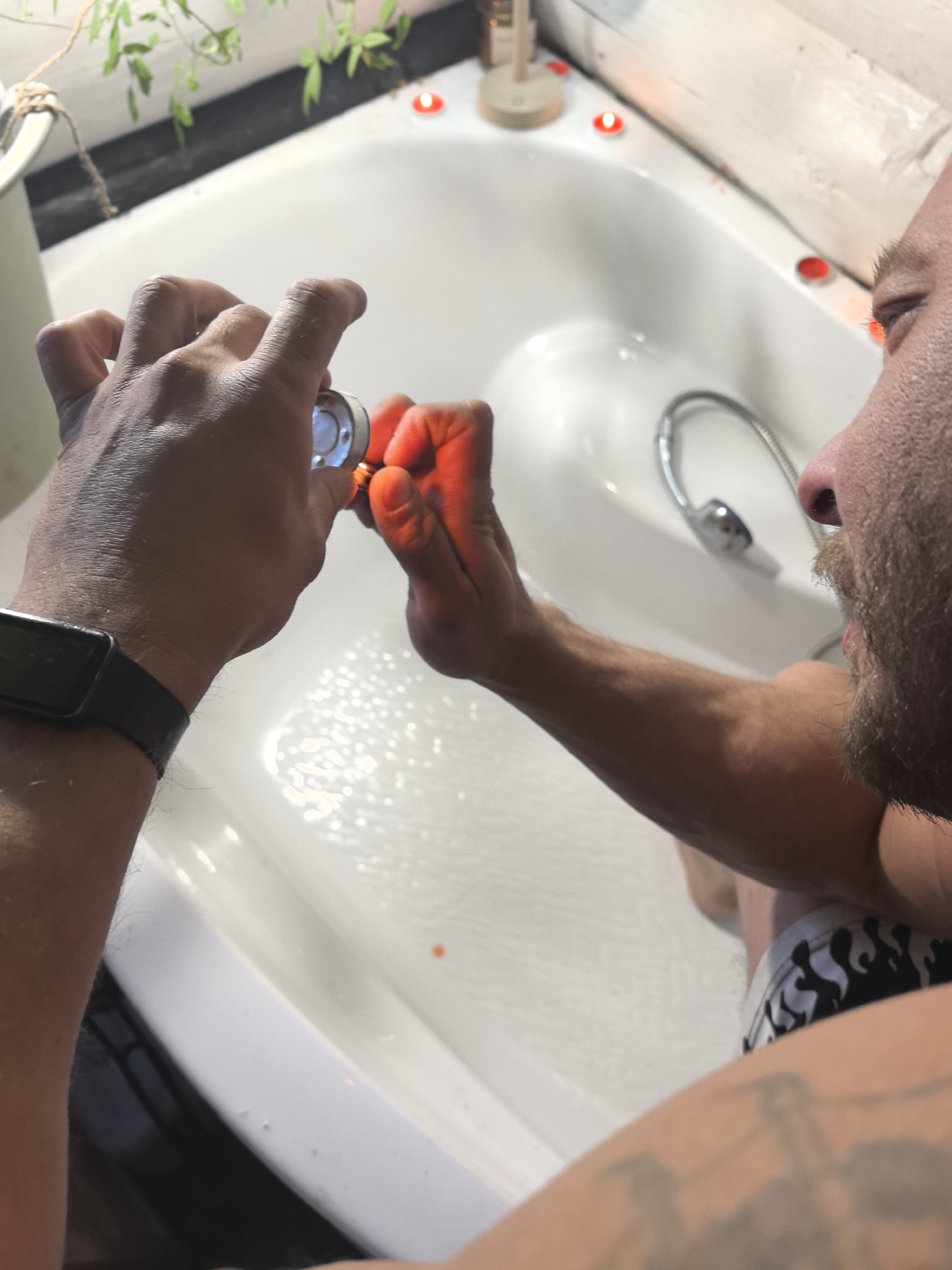 A man is depicted in a bathtub filled with water. He is holding a small, metallic object in his hands. He is using a lighter to heat it. The bathroom is decorated with small, red, candle-like lights along the edge of the bathtub. The location is Noviy, Russia.