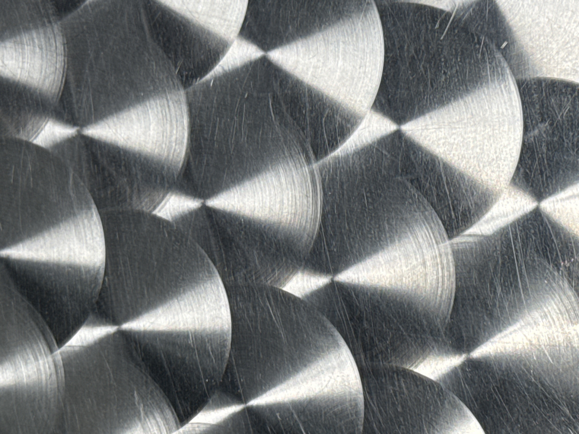 The image displays a close-up of a textured metal surface. The texture consists of overlapping circular patterns, each with a distinct center point and concentric rings emanating outwards. The surface appears to be brushed or scratched, exhibiting a non-uniform, directional pattern. The color is primarily silver or gray, with variations in tone suggesting reflections and shadows. The overlapping circles create geometric shapes, particularly diamond or triangular patterns, where the circles intersect.
There is no mention of the location in the city of Moscow, Russia in the image.