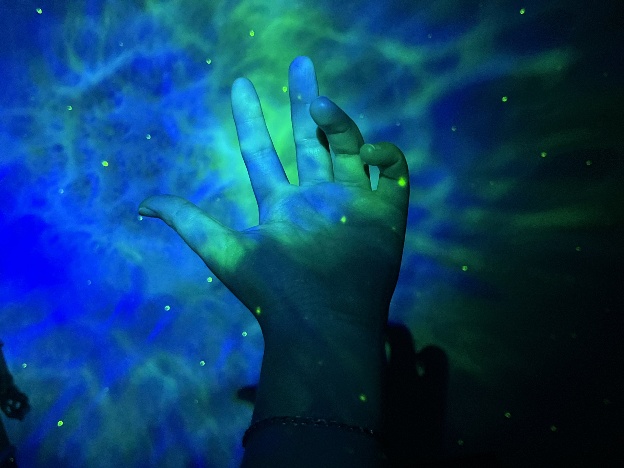 The image depicts a close-up of a person's hand, with fingers extended, against a background of blue and green swirling lights. The hand appears to be illuminated by the same colorful light, creating a glowing effect. Small green dots are scattered throughout the background, resembling stars or illuminated particles. The lighting suggests a dark environment, possibly a room with a projector or a light installation. The location in Saint Petersburg, Russia is not discernible from the image itself.