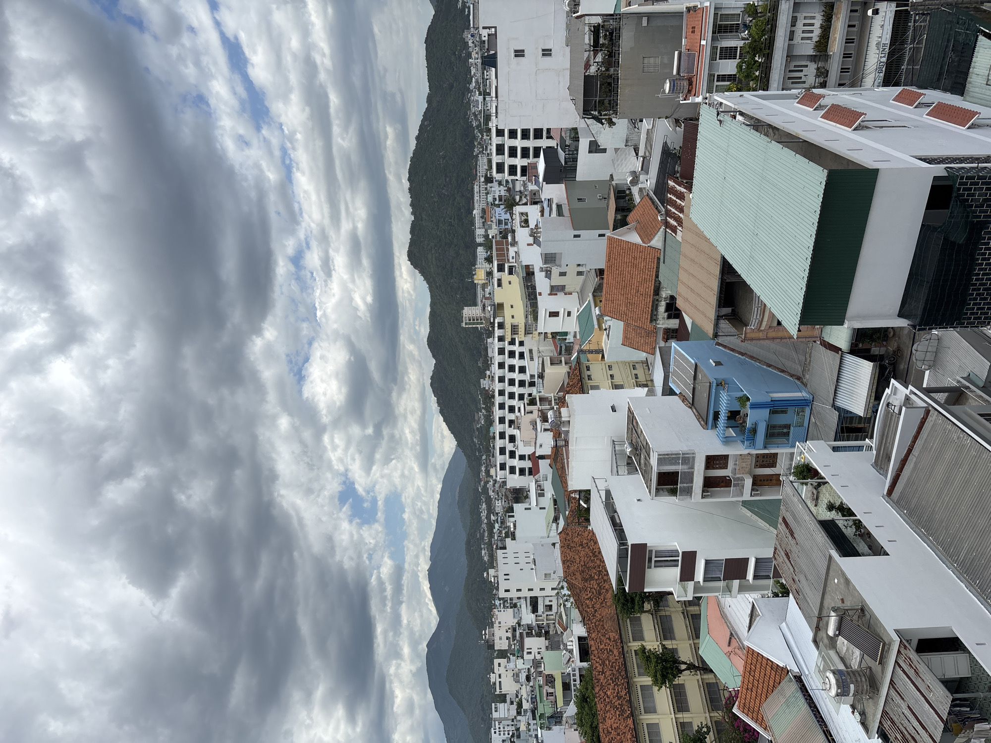 The image is a high-angle shot of a dense urban landscape in Nha Trang, Vietnam, under a cloudy sky. The foreground and midground are filled with tightly packed buildings of varying heights and architectural styles, characterized by flat roofs, numerous windows, and balconies. Many buildings are white or pastel-colored, with some featuring terracotta-tiled roofs and others with corrugated metal roofing. The background is dominated by lush, green hills that rise behind the city, suggesting a coastal or mountainous setting. The sky is overcast with thick, grey clouds, obscuring any direct sunlight and casting a diffused light over the scene. No people or specific actions are clearly visible, contributing to a sense of a static, observational view of the urban environment.