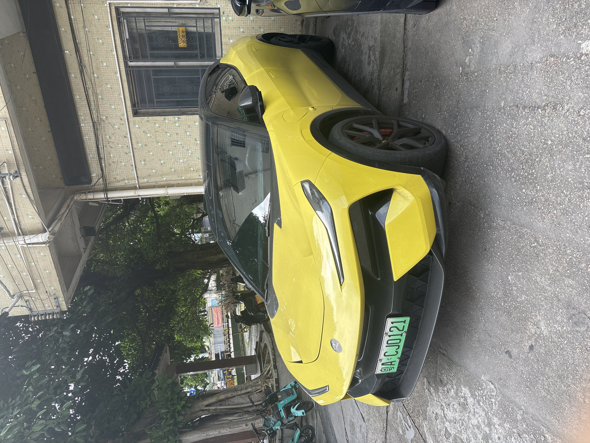 A bright yellow Lotus Eletre electric SUV is parked on an asphalt surface in front of a light yellow tiled building. The car has a black roof and black rims with red brake calipers. A license plate reading "A·CJ0121" is visible on the front of the car. To the left of the SUV, two teal electric scooters are parked. The building has several windows with metal security bars and what appears to be an air conditioning unit on the exterior. Overhead, electrical wires and conduit are visible, as well as the branches of a large tree. In the background, a construction fence is present, suggesting ongoing development. The scene appears to be located in the Nanhai District of China.
