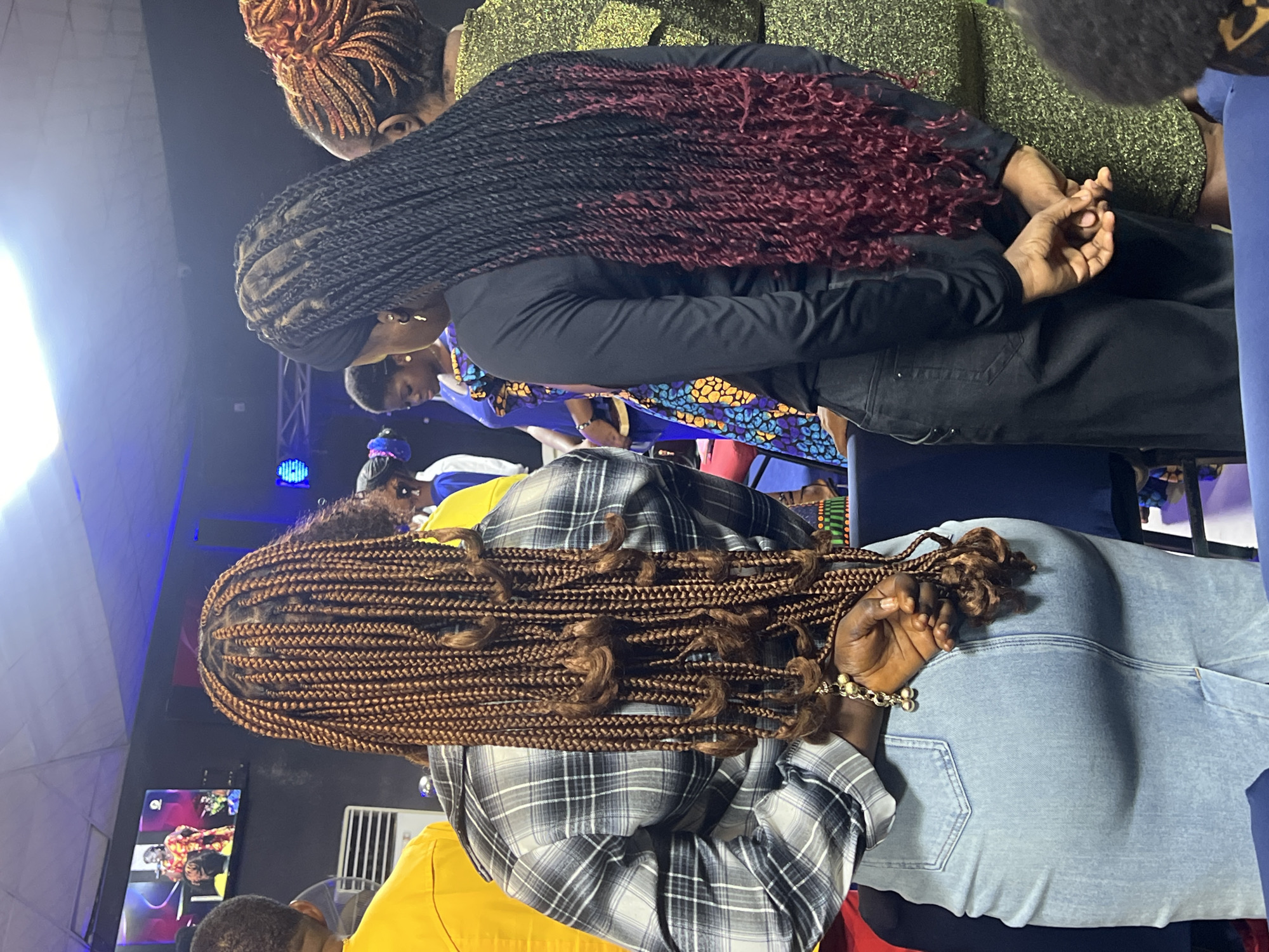 In Nkpor, Nigeria, a group of people are gathered indoors, likely at a church or event hall. The scene focuses on the backs of several women. In the foreground, a woman with long, reddish-brown box braids wears a grey and black plaid shirt and a light blue denim skirt. Her hands are clasped behind her back, adorned with a bracelet of gold-toned beads. To her right, another woman has long, black box braids that transition to a deep red at the tips. She wears a black long-sleeved shirt and black pants. Her hands are also clasped behind her. A third woman, seen from the side, wears a shimmering green garment. In the background, a large screen displays a performance by a woman in a vibrant orange and yellow outfit. The environment is dimly lit, with blue stage lighting visible in the distance.