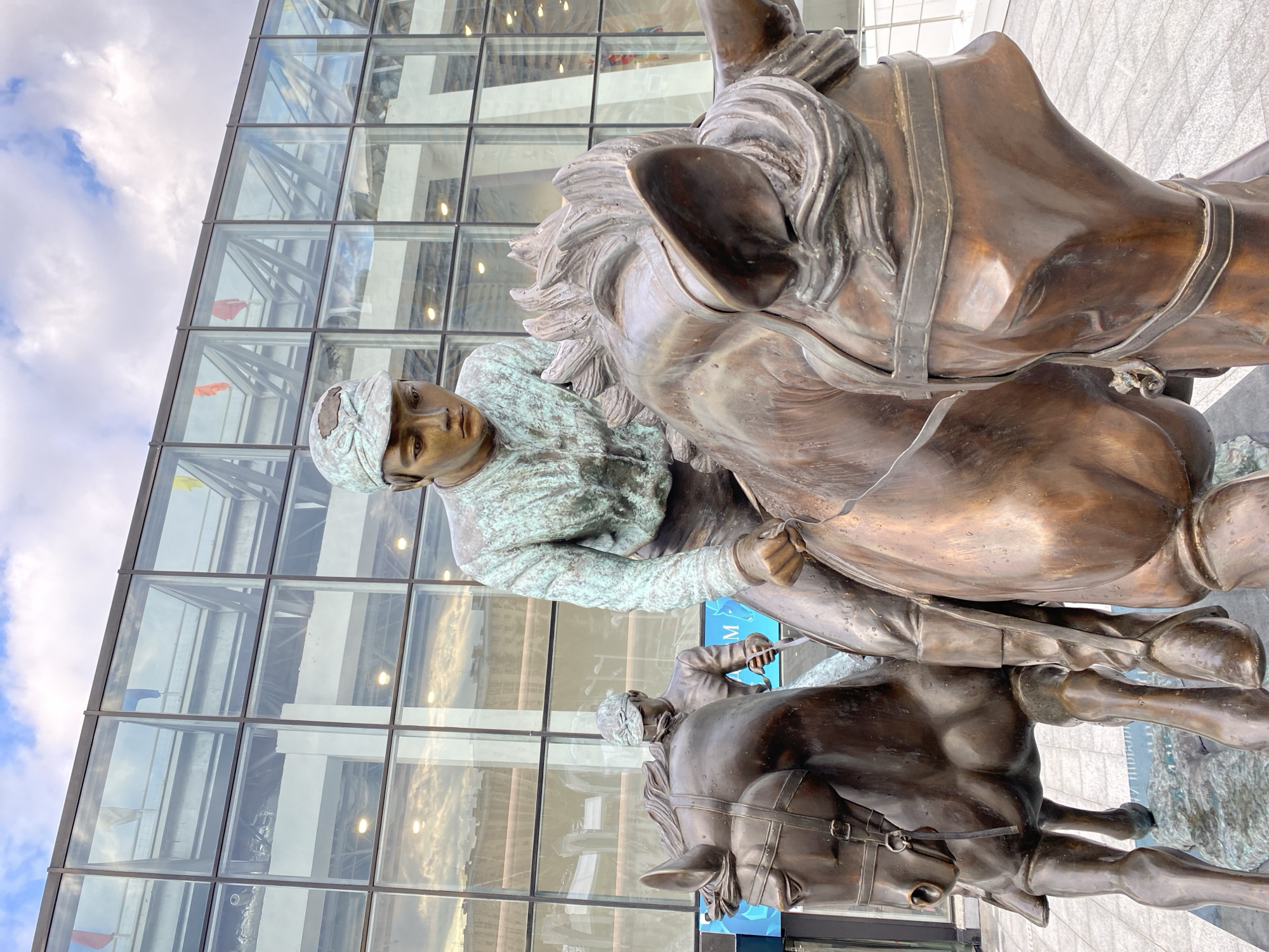 Two bronze statues of jockeys riding horses are positioned in front of a modern glass building. The jockey in the foreground, wearing a cap and a patterned shirt, is depicted in mid-action, holding reins. The horse he is riding is also rendered in bronze, with a bridle and saddle. Behind them, a second jockey on a horse is visible. The background features a large glass facade of a building with a cloudy sky overhead. The overall scene suggests an outdoor public space, potentially a park or plaza, in the city of Kazan, Russia.