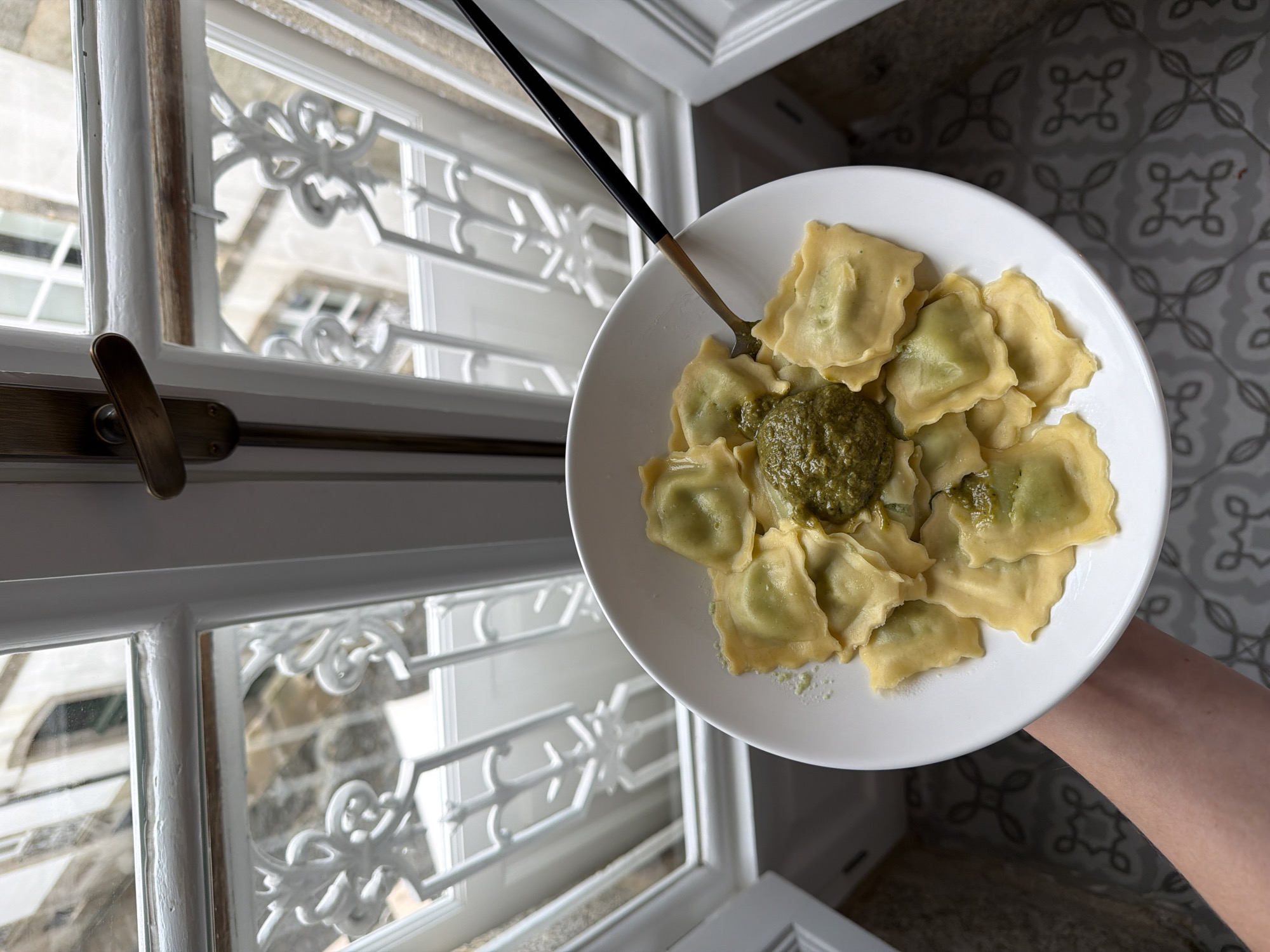 Here is a detailed description of the image:
**Scene:** An eye-level, slightly angled, close-up shot shows a plate of ravioli with pesto sauce next to a window. A hand holds the plate.
**Location:** Based on the window style and the floor tiling pattern, it is possible that this photo was taken indoors at a building in Santiago De Compostela, Spain.
**Objects:**
* A white plate filled with about a dozen ravioli pasta.
* A scoop of green pesto sauce sits in the center of the ravioli.
* A fork, with a black handle and a golden tip, rests on the plate.
* A multi-pane window with decorative white ironwork framing the glass. The window is partially open.
* A metal handle or latch is visible on the window frame.
* A light-colored tiled floor.
**Environment:**
The lighting appears to be natural daylight coming through the window. The overall color scheme is light and bright with white, yellow, and green tones. The background seen through the window seems to be a stone building.
**People:**
Only a portion of a person's arm and hand is visible, holding the plate of food.
**Actions:**
The act of presenting a plate of food.
**Interactions:**
The hand interacts with the plate.