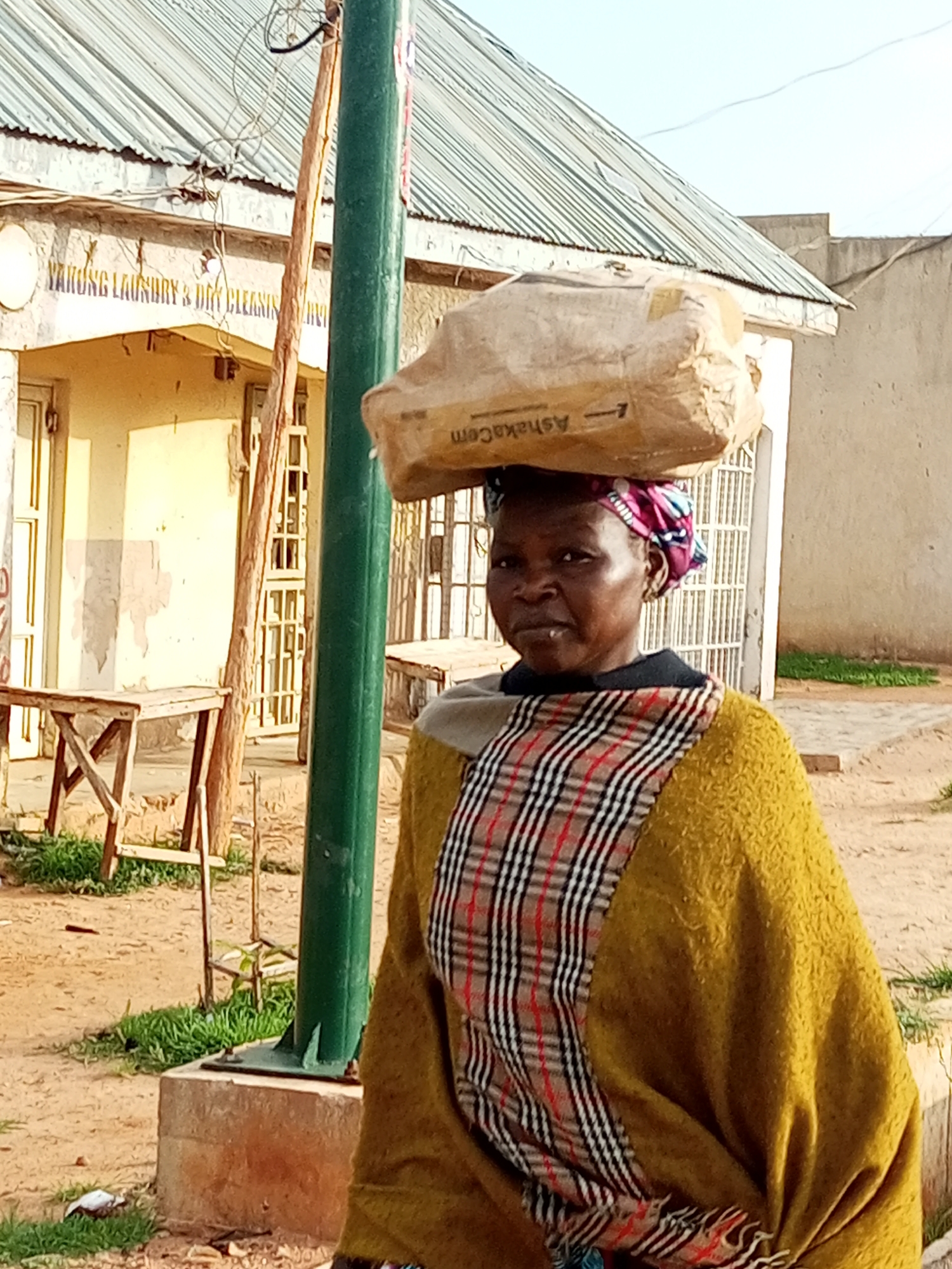 The image captures an outdoor scene in the city of Gombe, Nigeria. A woman is the central subject, carrying a large, wrapped package on her head. She is facing the camera and appears to be standing on a dirt or sand path. Behind her, a building with a corrugated metal roof is visible. The building has a sign that partially reads "ARUNG LAUNDRY & DRY CLEANING". There are also some wooden tables and chairs set up in front of the building. A tall, green pole stands to the left of the woman, and electrical wires are strung overhead. The overall environment suggests a commercial or residential area within the city.