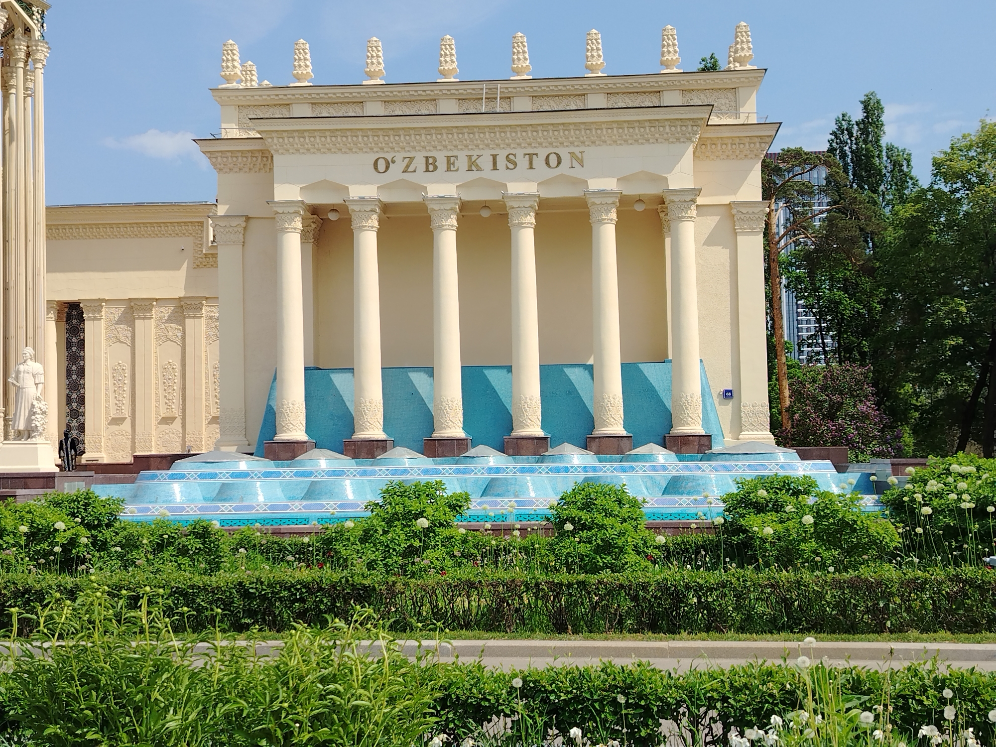 The image shows the Uzbekistan Pavilion in VDNH (Exhibition of Achievements of National Economy), located in Moscow, Russia. The pavilion is a beige-colored structure with a classical design, featuring a facade adorned with columns and topped with decorative elements. The name "O'ZBEKISTON" is inscribed above the columns. Below the colonnade, there is a raised platform with blue tiled surfaces. The pavilion is surrounded by greenery, including hedges, bushes, and trees. The weather appears to be sunny, with a clear blue sky visible in the background. There is a statue to the left of the building.