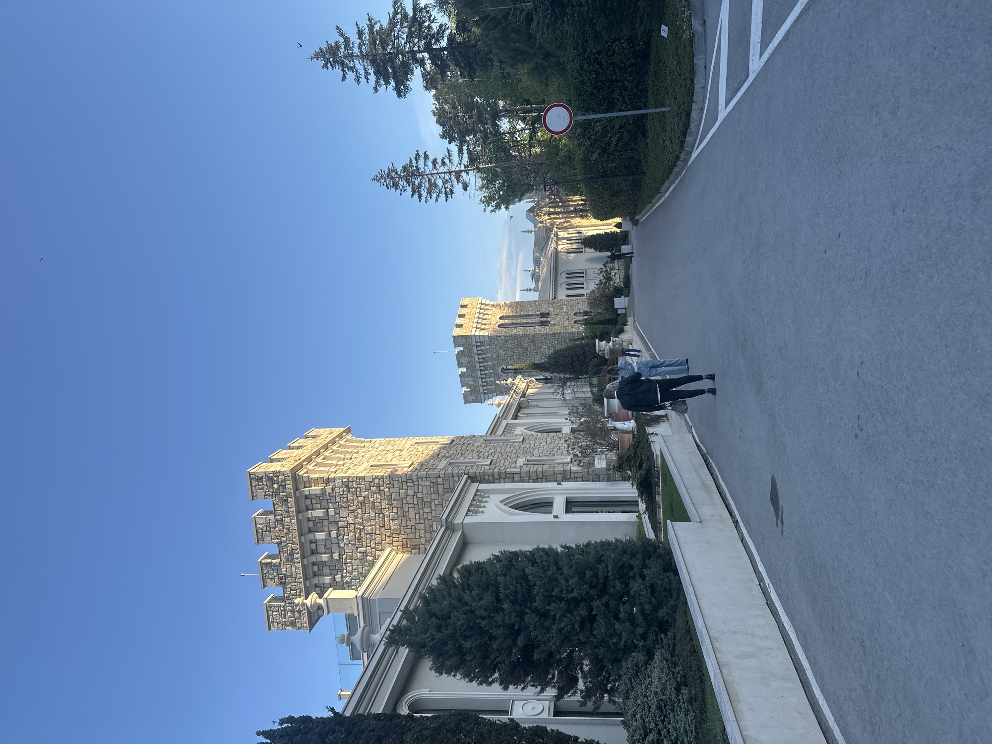 An eye-level, medium-shot photograph shows Euxinograd palace, located in the city of Varna, Bulgaria. The image is vertically oriented. The sky is clear and blue. The palace is built with stone that appears tan or beige in color. The architecture of the palace is ornate with multiple tiers, arches, and details. A paved street is alongside the palace. Two individuals are walking on the street. There are trees and shrubs located alongside the palace. A no-entry sign is posted on the side of the road.