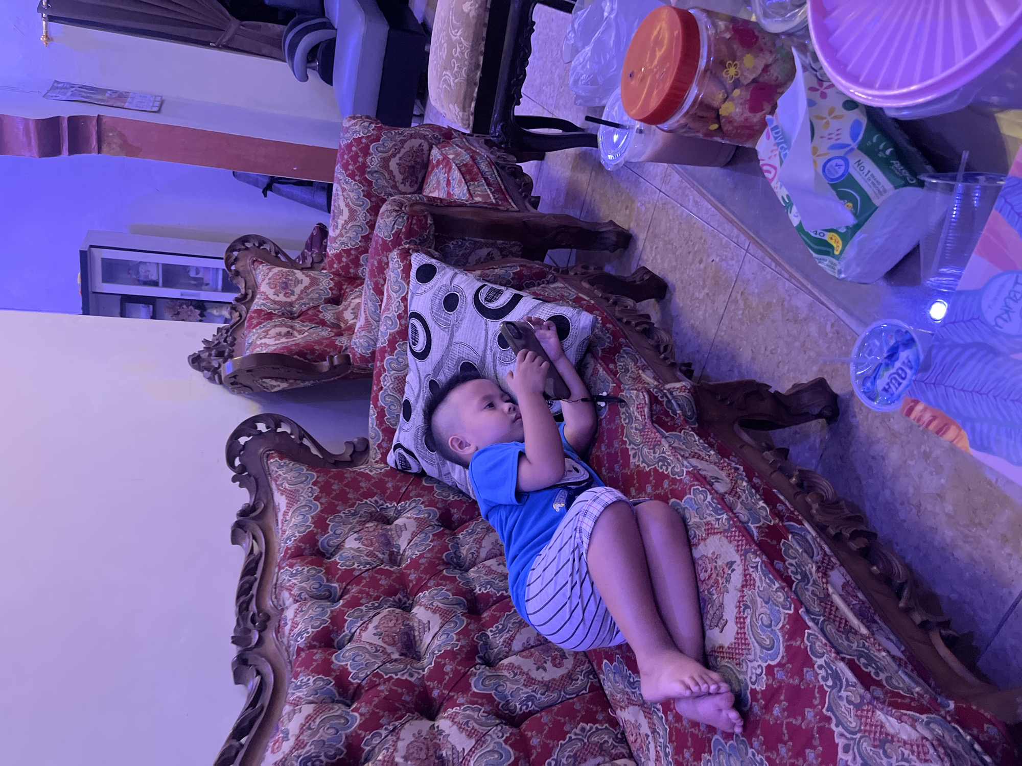 A young boy lies on his side on a patterned sofa, looking at a phone. He is wearing a blue t-shirt and striped shorts. The sofa is ornate with carved wooden details and a red and gold floral pattern. A patterned pillow is under his head. In the background, another armchair with similar upholstery is visible, as well as a cabinet. On a glass-top table in the foreground, there are drinks, a jar of cookies, and tissues. The lighting is dim with a purple hue. The location is a residential interior in Jatisari, Indonesia.