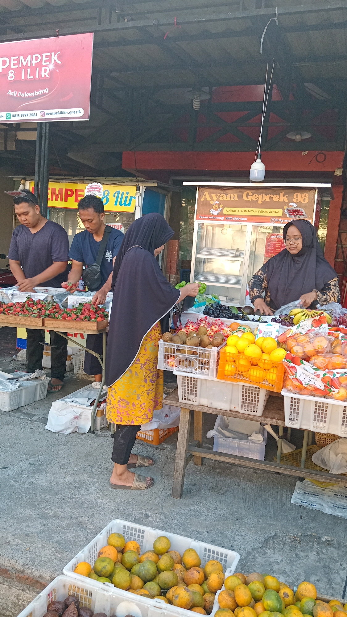 In Randuagung, Indonesia, a street vendor stall displays an assortment of fruits. Three individuals are present: two men standing on the left, attending to a crate of strawberries on a wooden table, and a woman in the center wearing a yellow patterned dress and a black hijab, interacting with a female vendor behind the fruit stall on the right. The woman in the yellow dress appears to be selecting green grapes from a crate. The fruit stall features kiwis, oranges, bananas, grapes, and other citrus fruits displayed in plastic crates. Above the fruit stall, a sign for "Ayam Geprek 98" is visible. The scene is set outdoors, with a concrete ground surface and the stall structure suggesting a permanent or semi-permanent market setup. The lighting indicates it is daytime.