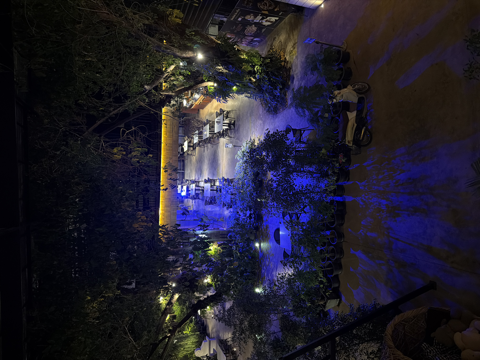This image depicts an outdoor seating area of a restaurant or cafe at night. The scene is illuminated by artificial lights, including string lights in the trees and a prominent yellow light fixture on a building in the background. The area is furnished with multiple tables and chairs, suggesting it's a place for dining or socializing. Lush greenery, including trees and potted plants, surrounds the seating area, creating an inviting and natural ambiance.
A blue light casts dramatic shadows on the ground and walls, adding a theatrical element to the night scene. In the middle ground, a scooter is parked, and a person is seated on it, partially obscured by shadows and foliage. The overall atmosphere is serene and perhaps a bit mysterious due to the nighttime lighting. The presence of trees and outdoor seating suggests a warm climate.
There are some signs visible on the building in the background, but the text is not clearly legible. The image conveys a sense of a peaceful evening in an outdoor dining establishment. The specific location context provided is Ban Phonsavang Neua, Laos, which aligns with the tropical-like vegetation and outdoor dining setup.