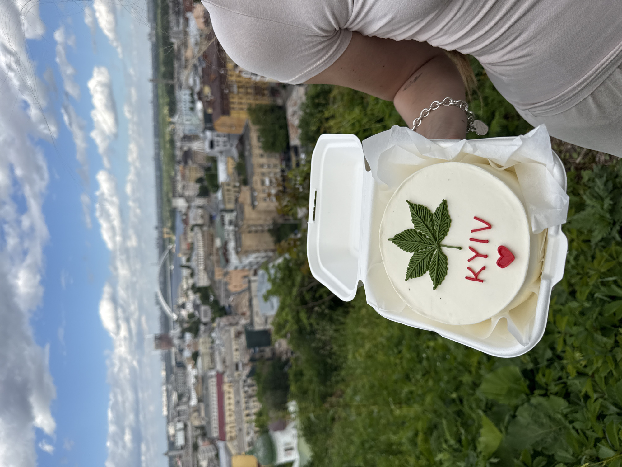 The image shows a person holding a white takeout container with a small round cake inside. The cake is decorated with white frosting, a green cannabis leaf, and the word "KYIV" written in red icing with a small red heart. The person is wearing a light-colored short-sleeved shirt and a silver bracelet.
In the background, there is a cityscape with a large pedestrian bridge crossing a wide river, the Dnipro. The city is characterized by numerous buildings and green spaces. The sky is blue with scattered white clouds. The location appears to be a viewpoint overlooking Kyiv, Ukraine, likely on a hillside or elevated area. The presence of the cake with "KYIV" on it further confirms the location. There is also vegetation near the person, suggesting a grassy or park-like setting.