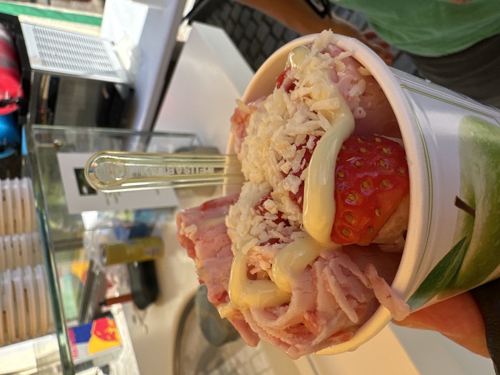 A close-up, eye-level shot shows a person holding a paper cup filled with rolled ice cream. The ice cream is pink and appears to be flavored with strawberries, as evidenced by a whole strawberry on top. It is drizzled with a white, creamy sauce and sprinkled with shredded white pieces, possibly coconut or white chocolate. A translucent yellow plastic utensil is stuck into the ice cream. The cup has a green apple design on it. In the background, a counter is visible with various items, including a red and yellow Red Bull can, a yellow can, and a glass enclosure containing what appears to be a slushy machine. The setting seems to be an outdoor kiosk or stand. The general location is identified as Schönau am Königssee, Germany.