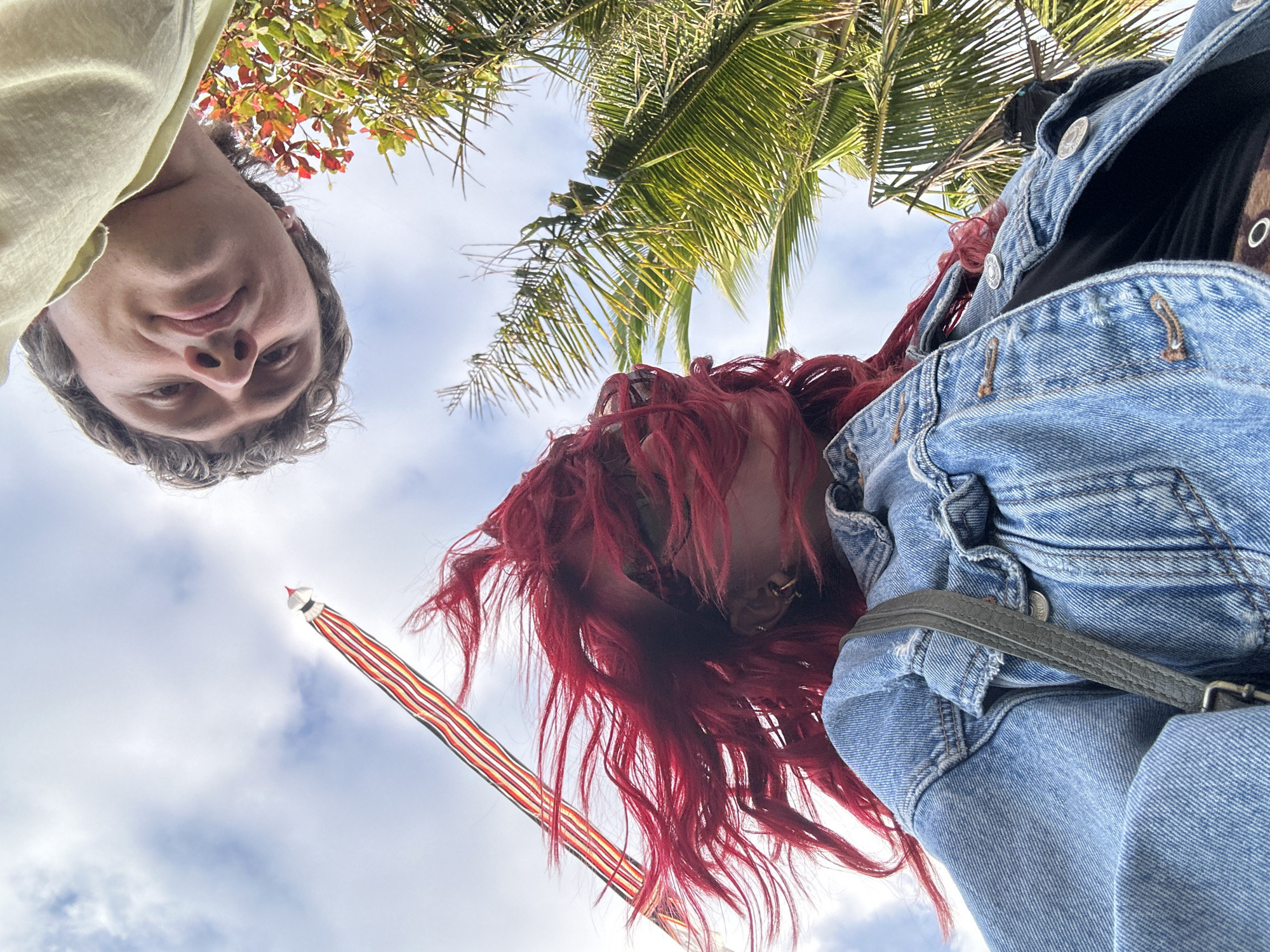 In Mengwi, Indonesia, two individuals are captured from a low-angle perspective, looking upwards. A person with vibrant red hair, wearing a denim jacket and a dark top, is in the foreground. Their hair is dramatically windblown, obscuring much of their face. To their right, a person wearing a pale yellow t-shirt is partially visible, their face turned towards the camera with a relaxed expression. Above them, a tall, striped kite with red, yellow, and black bands is flying against a backdrop of a cloudy sky. Palm fronds are visible on the right side of the frame, indicating a tropical environment. The image is a self-taken photograph, suggesting a casual outdoor setting.
