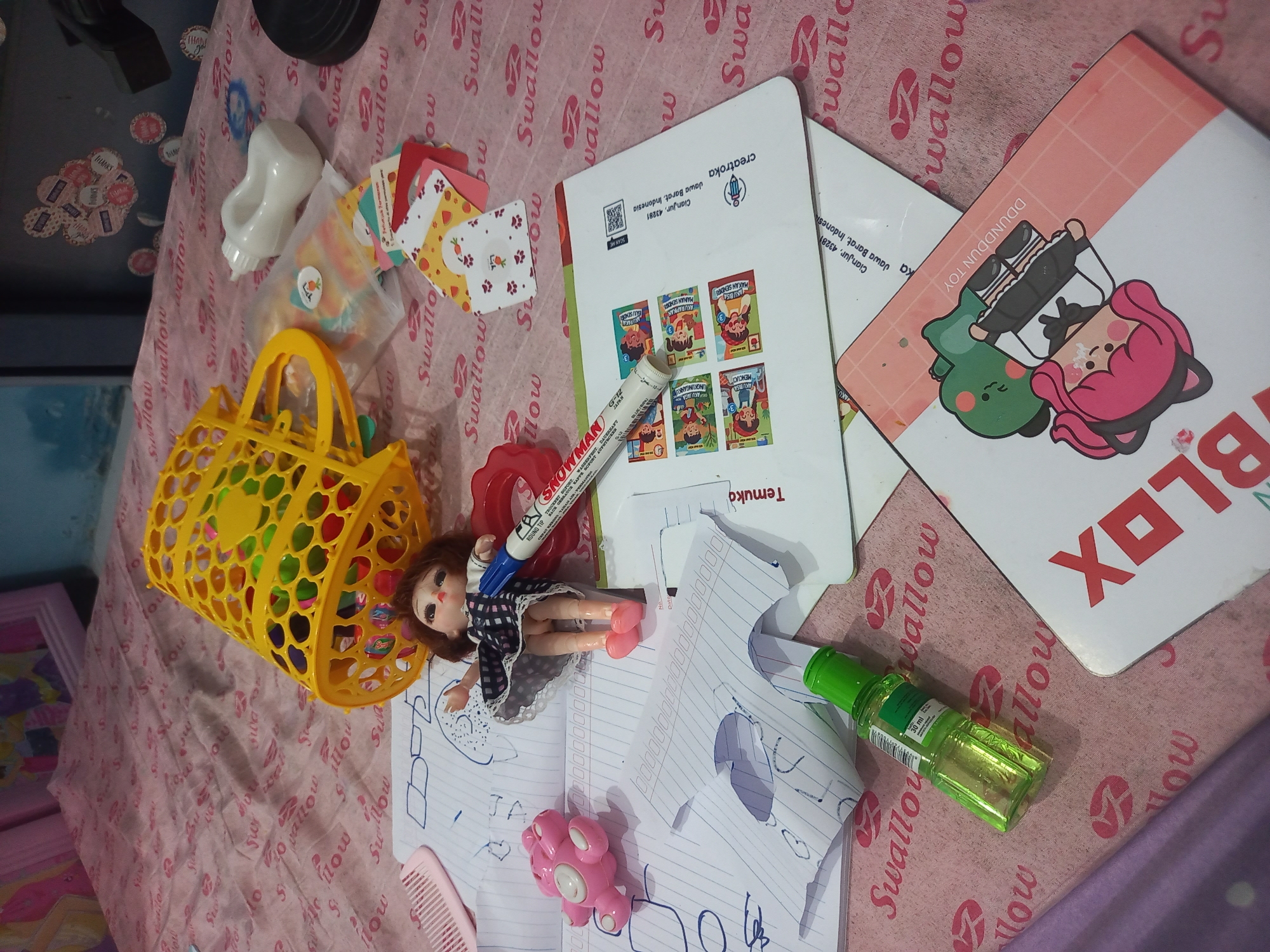 The image depicts a still life of various objects scattered on a pink, patterned fabric. In the foreground, a small, green bottle with a label is visible, along with pieces of torn paper. A white card with red lettering, possibly "BLOXX," is also present. Further back, a doll with brown hair, wearing a black and white checkered dress and pink shoes, lies on more crumpled paper. A blue marker with the brand "SNOWMAN" is positioned near the doll. To the left, a pink, toy-like object resembling a bear is present. In the upper portion of the image, a yellow, perforated basket is overturned, containing colorful objects. A white baby bottle is also visible near the basket. The background shows a blurred pink and white patterned surface. The scene appears to be indoors, with a hint of a framed picture or artwork in the far upper left corner. The image shows the location of "Cianjur, Jawa Barat, Indonesia" on one of the cards, indicating the origin of some of the items.