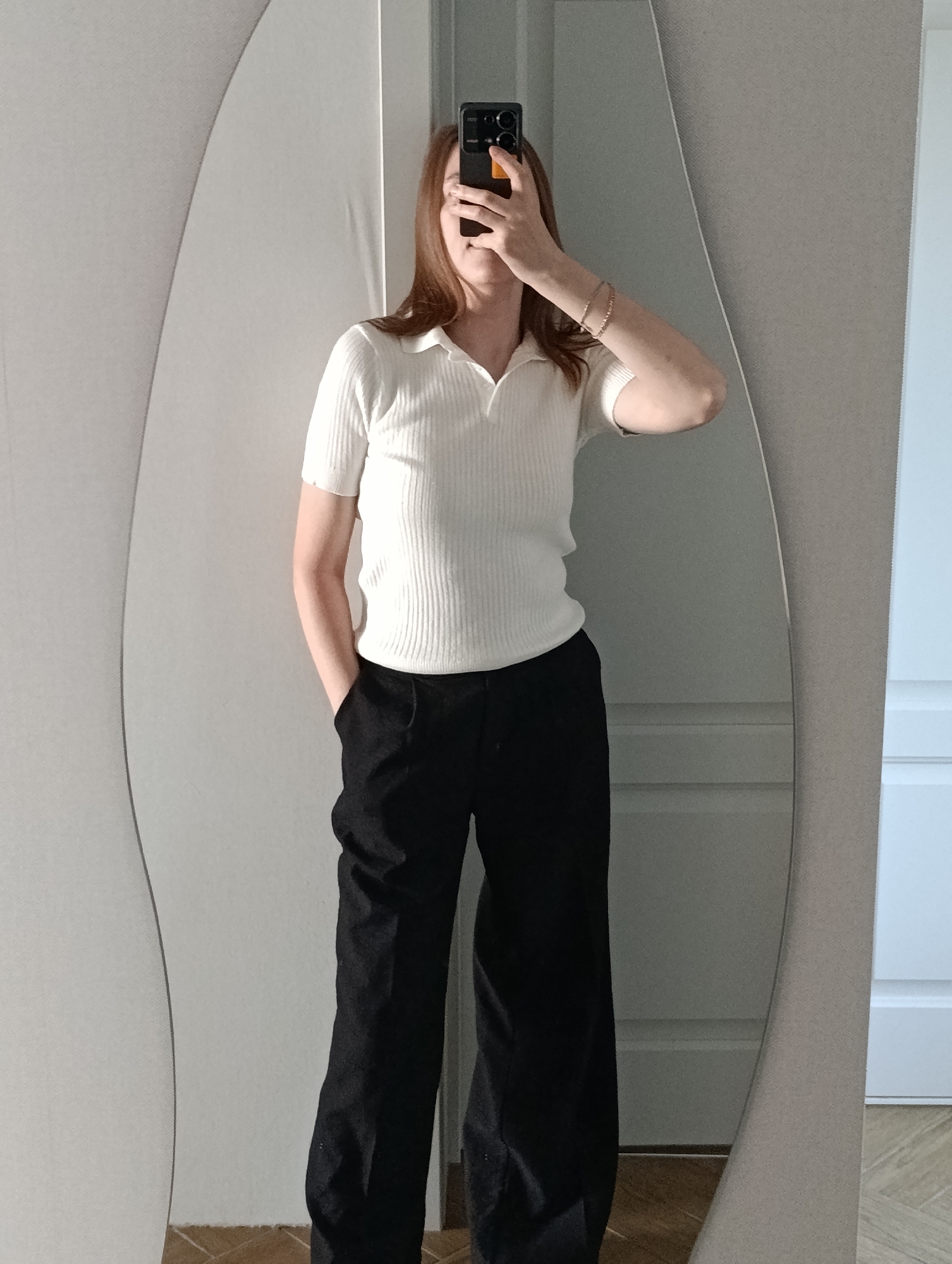 A person is taking a mirror selfie indoors. The individual is wearing a white ribbed polo shirt and black wide-leg pants, with one hand in a pocket and the other holding a smartphone up to their face. They are also wearing what appears to be bracelets on their right wrist. The mirror has an irregular, curved shape. The background shows a light-colored wall, part of a door, and wood flooring. The specific location in the city of Chaikovsky, Russia, cannot be determined from the image.