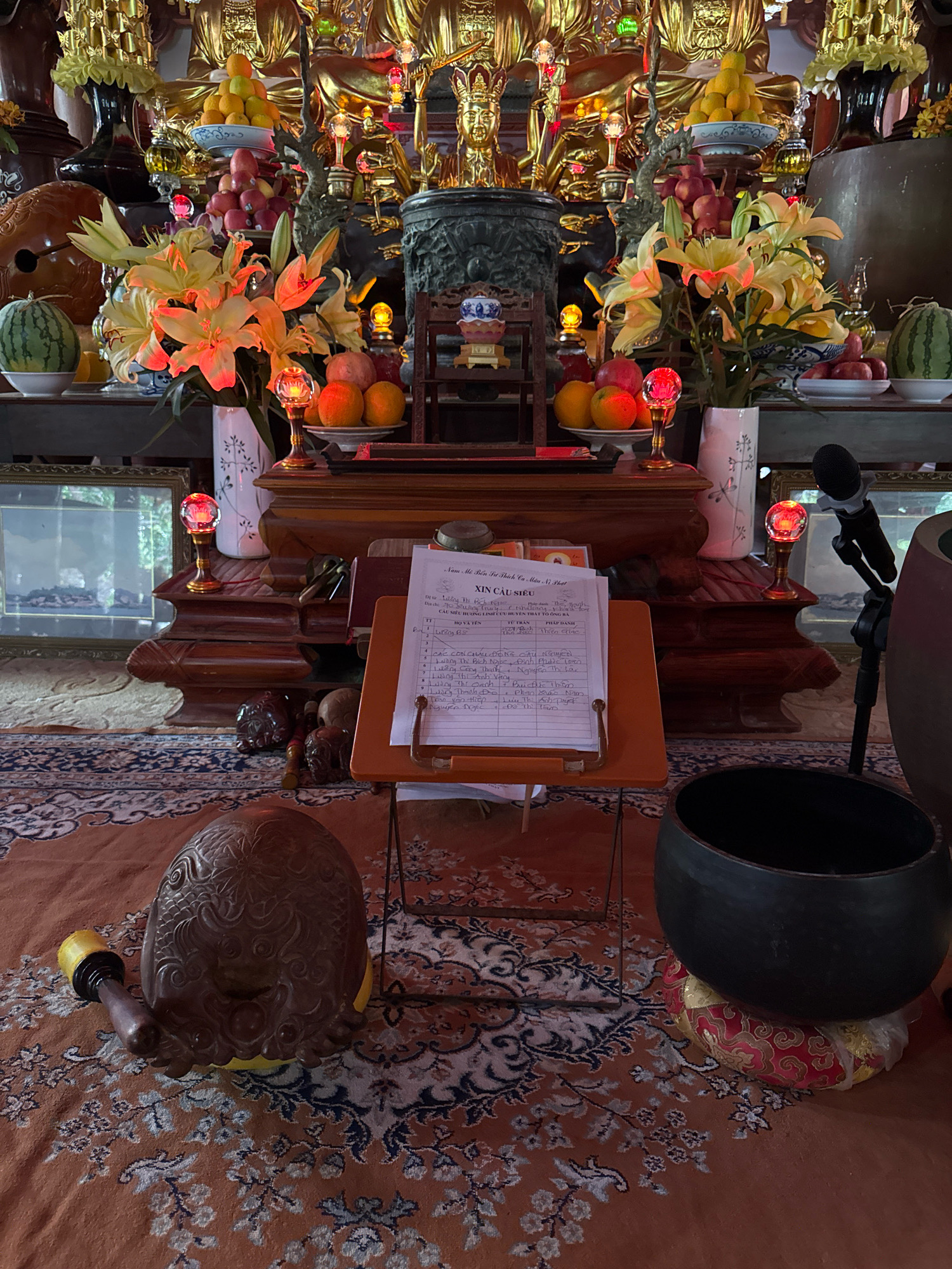 This image captures a close-up view of an altar within a Buddhist temple or shrine, likely located in Nha Trang, Vietnam. The altar is adorned with offerings of fruit, including oranges and what appear to be apples, as well as a bouquet of lilies. Decorative elements such as golden statues of deities, ornate vases, and illuminated red glass orbs are visible in the background and on the altar itself. In the foreground, a large, dark metal bowl rests on a patterned cushion. To the left of the bowl is a carved wooden object with a mallet. A microphone stands to the right of the altar, suggesting a service or presentation is taking place or has recently concluded. A sheet of paper with handwritten text, likely a prayer or list, is propped up on a small stand in front of the altar. The flooring is covered by an intricately patterned rug with dominant shades of orange and blue. The overall scene suggests a reverent and ceremonial setting.