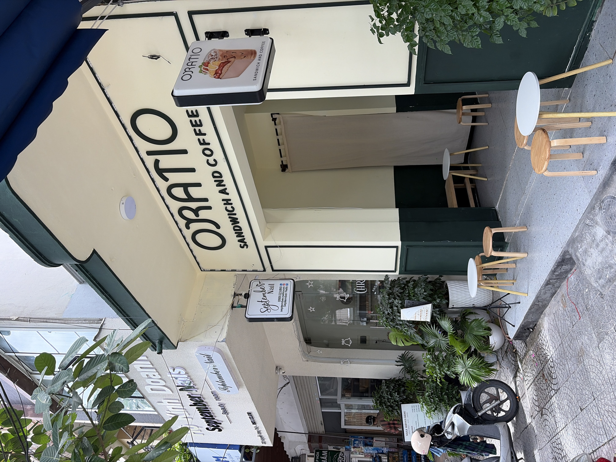 This image shows the exterior of two businesses on a city street in Da Nang, Vietnam. On the right is "ORATIO", a sandwich and coffee shop. The shop's name is in large, dark green, sans-serif font on a pale yellow facade, with a smaller sign depicting a sandwich and iced coffee. There are two small, light-colored round tables with wooden stools outside the entrance, and a beige curtain hangs inside the doorway. To the left is "September Nail", a nail salon. Its name is in a similar font, with a smaller "Beauty room" below it. A hanging sign for "September Nail" provides a phone number and social media handles. A scooter with a helmet parked beside it is in front of the nail salon, along with potted plants. The street is paved with patterned tiles, and some of the buildings have trees and foliage growing on them. The overall impression is of a well-maintained, modern commercial streetscape.