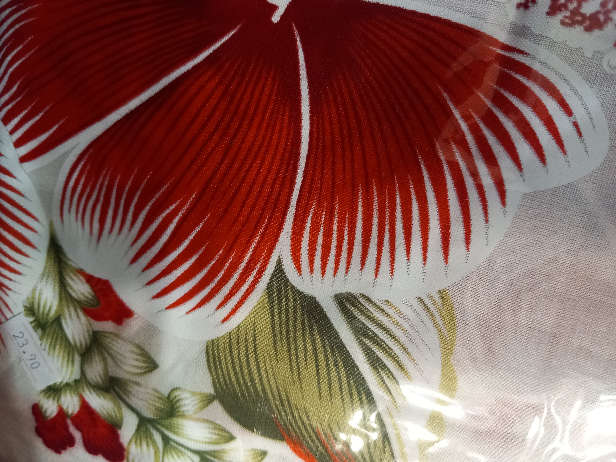 An indoor, close-up shot shows a piece of fabric with a floral pattern. A large red and white flower petal dominates the right side of the image, with fine red lines radiating outward on a white background. To the left of the petal, a cluster of green leaves and red flower buds are visible. A small white price tag is affixed to the fabric in the upper left quadrant, displaying the numbers "23.90" upside down. The fabric appears to be covered in a clear plastic wrapping, which causes some glare and distortion in the image. The setting is not specified beyond the general location of "Unknown, Unknown."