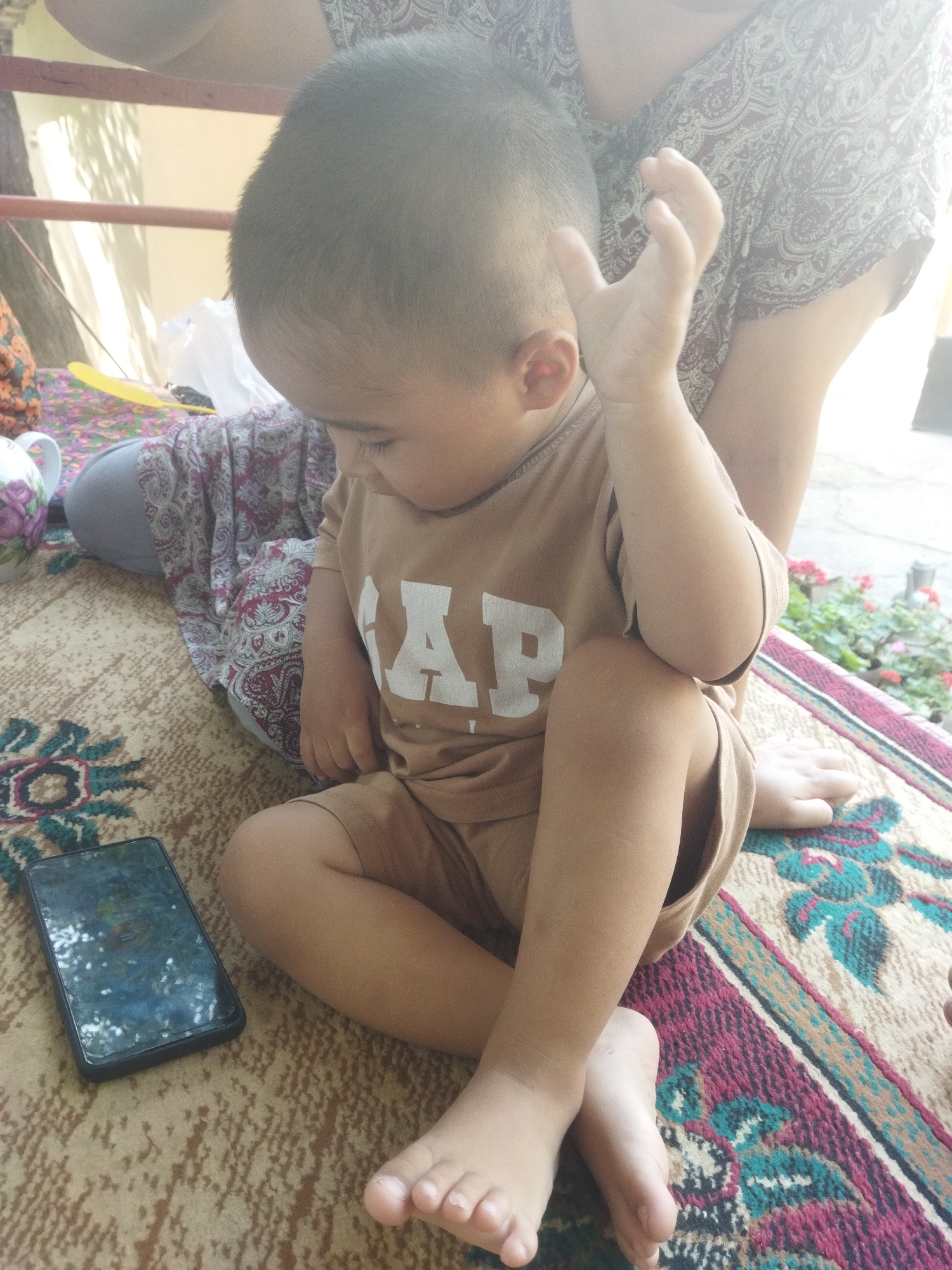 In Mailuu-Suu City, Kyrgyzstan, a young child is sitting on a carpeted surface. The child is wearing a tan t-shirt with the letters "GAP" in white. The child's left hand is raised, and the child is looking down. A smartphone is lying on the carpet to the child's left. A person, likely an adult, is partially visible in the background, wearing a patterned garment. The environment appears to be outdoors, with some greenery and a structure that could be a fence or railing visible in the background.