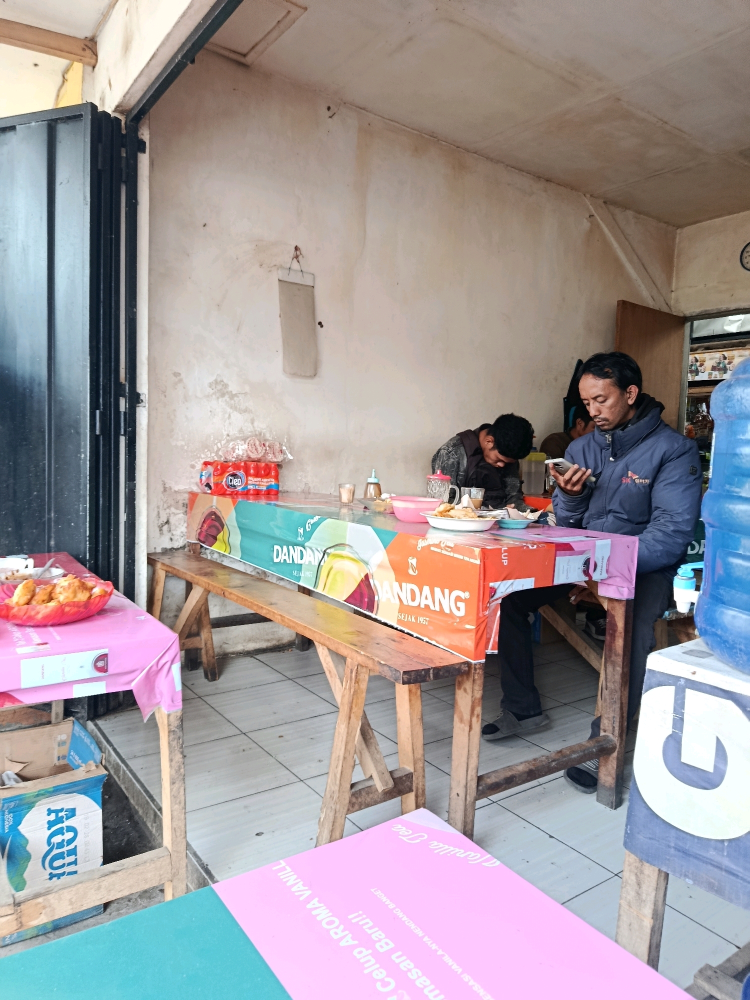 The image depicts an outdoor food stall located in Bululawang, Indonesia. In the foreground, a man in a dark blue jacket is seated at a table, engrossed in his mobile phone. He is positioned on the right side of the frame, with a large blue water dispenser to his right. Across from him, another person, partially obscured and appearing to be younger, is also seated at the table, looking down, possibly eating.
The table is covered with various items including plates of food, drinks, and some packaging. The table itself is draped with colorful banners. One banner, prominently featuring the word "DANDANG" and "SEJAK 1957," appears to be advertising a food product. To the left of the main table, another table is set with a pink tablecloth, holding a bowl of food.
Behind the tables, a wooden bench is visible, with several red and white packaged drinks stacked on top. The stall appears to be set up along a walkway or street, with a building structure behind it, characterized by light-colored, somewhat weathered walls. A metal gate or door is partially visible on the far left. The ground is tiled. The overall scene suggests a casual, perhaps local eatery.