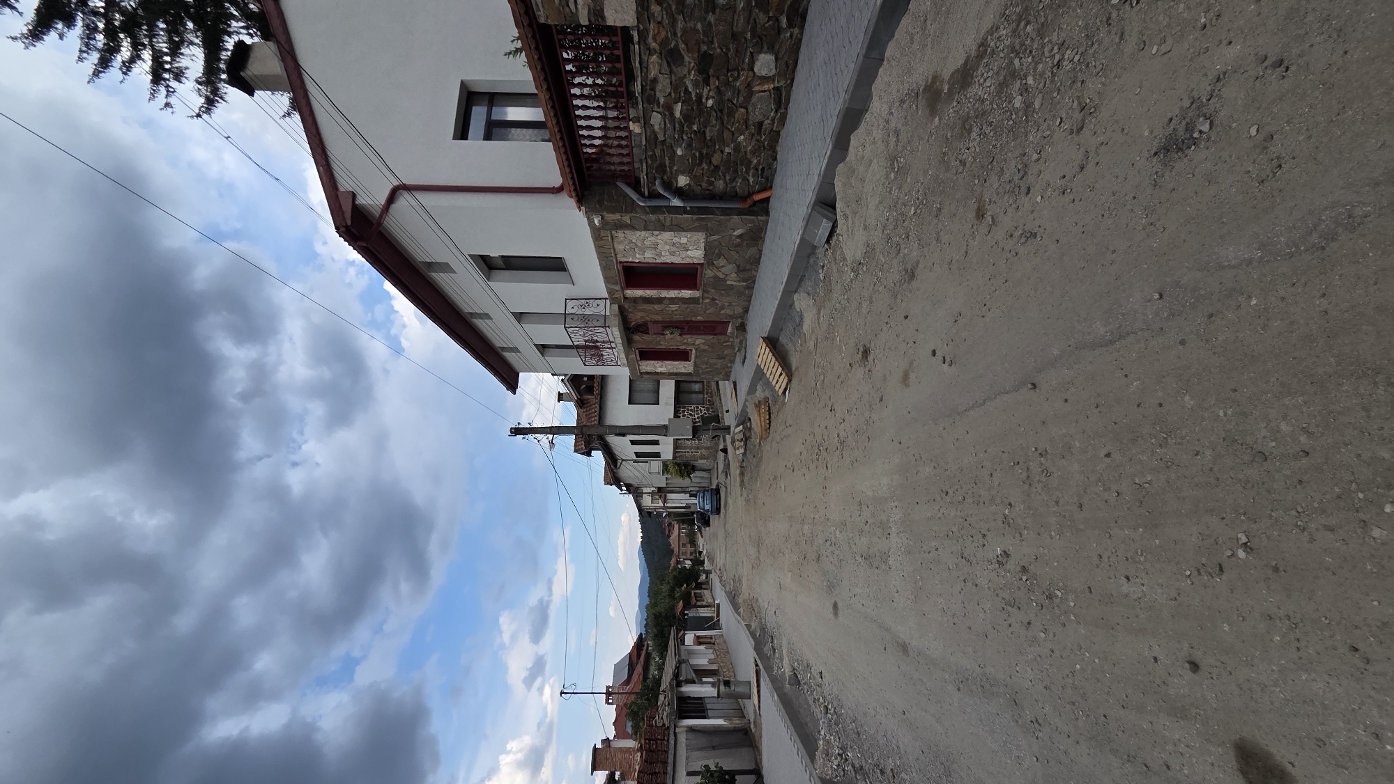The image depicts a street in the city of Bania, Bulgaria, that appears to be under construction or repair. The foreground is dominated by a dirt road, uneven and rough, with loose gravel and stones scattered across its surface. To the right of the road, a newly paved sidewalk or curb made of interlocking grey stones is visible, with piles of sand and construction debris nearby.
Along the sides of the street, traditional-looking houses with red-tiled roofs and white or light-colored facades line the way. Some of these houses have stone foundations and decorative elements, such as iron balconies. Red-painted window frames are visible on some of the buildings. Utility poles with electrical wires are present along the street.
In the background, the street recedes, with more houses and buildings visible, as well as some trees and rolling hills or mountains in the distance. The sky is partly cloudy, with dramatic grey clouds interspersed with patches of blue sky. A single blue car is parked on the side of the street, and several wooden pallets are stacked near the curb. The overall impression is a village street in the midst of renovation, with a mix of old architecture and ongoing infrastructure work.