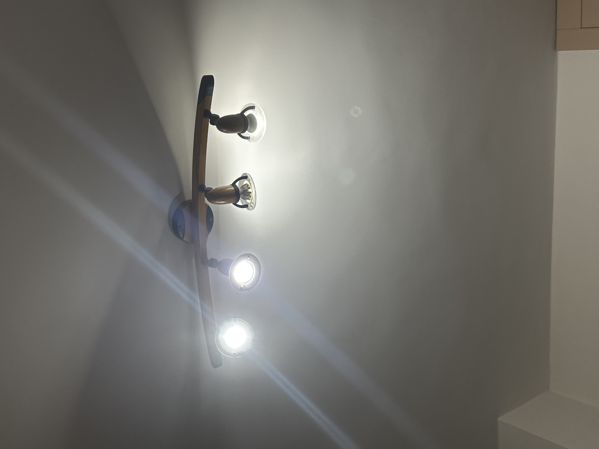 An indoor scene shows a wooden track light fixture mounted on a white ceiling. The fixture has four adjustable spotlight heads, each with a wooden casing and a clear glass lens. The lights are on, casting bright beams and lens flares onto the ceiling. The overall lighting is dim, with the ceiling becoming darker as it recedes from the lights. The lower portion of the image reveals a sliver of a white wall and a portion of a light-colored cabinet. The location is described as Unknown, Unknown.