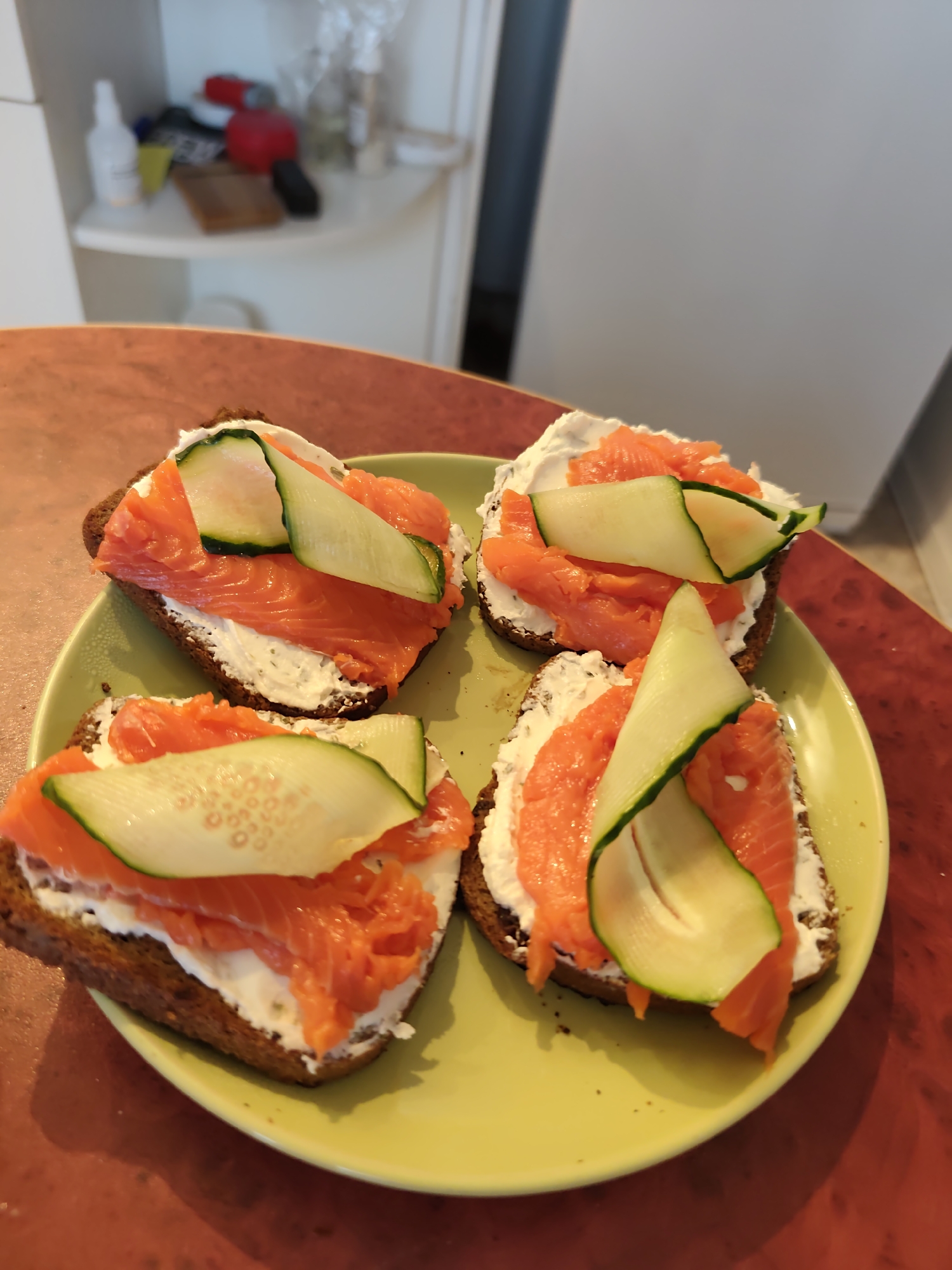 Four open-faced sandwiches are arranged on a light green plate, which sits on a reddish-brown table. Each sandwich consists of a slice of dark bread topped with a layer of white cream cheese, several slices of bright orange smoked salmon, and a curved slice of light green cucumber. The background shows a shelf with various items, including bottles and containers. This scene is indoors, possibly in a kitchen or dining area. No people are visible in the image. The context of Chaikovsky, Russia is not discernable from the visual information provided.
