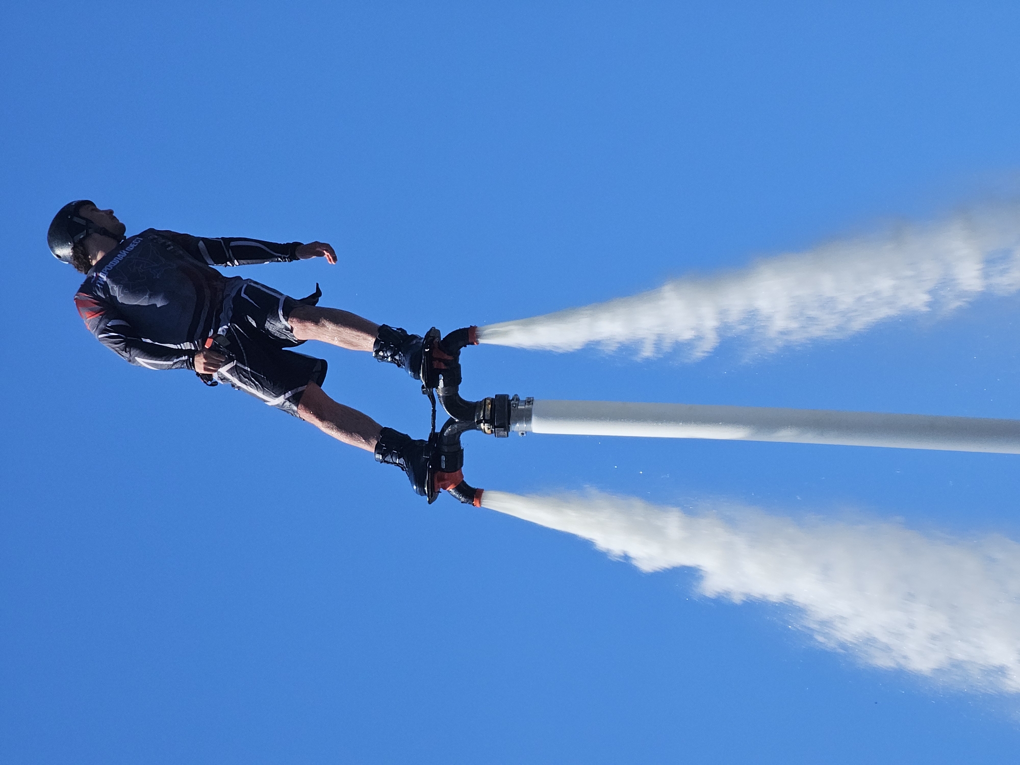 A person wearing a helmet, a dark long-sleeved shirt with red accents, and black shorts is suspended in the air against a clear blue sky, propelled by a white device with two red nozzles emitting powerful jets of water. The person is standing on top of this device, which appears to be a flyboard or a similar water-powered jetpack. The person is looking to the side, and their arms are slightly extended for balance. The water jets are creating a cloud of mist around the device. The overall scene suggests an outdoor water sports activity. There is no indication of the location being Moscow, Russia.