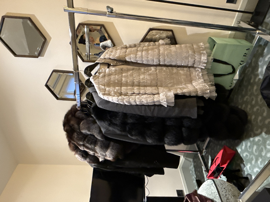 A clothing rack holds various garments. From top to bottom, there's a white, quilted jacket, a gray coat, a black fur coat, and another black coat. A light green shopping bag is situated near the jackets. Hexagonal mirrors decorate the wall above the rack. A television and other items are visible below the clothing. The room's decor features a patterned carpet.
