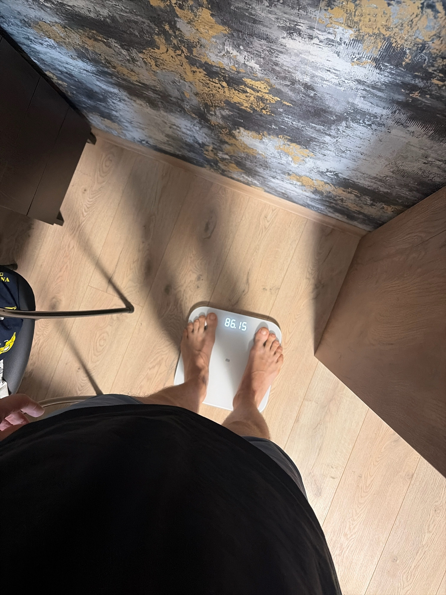 A person is standing on a white digital scale, which displays the weight "86.15". The scale is on a light-colored wooden floor. To the left of the scale, a portion of a black t-shirt is visible, and further to the left, a black chair with a dark fabric seat is partially in frame, with a glimpse of a dark garment with a yellow graphic on it. In the background, a wall features a textured wallpaper with a pattern of grey, black, and gold abstract strokes. To the right of the scale, a wooden cabinet or piece of furniture is visible. The lighting suggests an indoor environment, likely a home, and the scene is captured from a high-angle perspective looking down. There is no information to determine the location in Yekaterinburg, Russia.