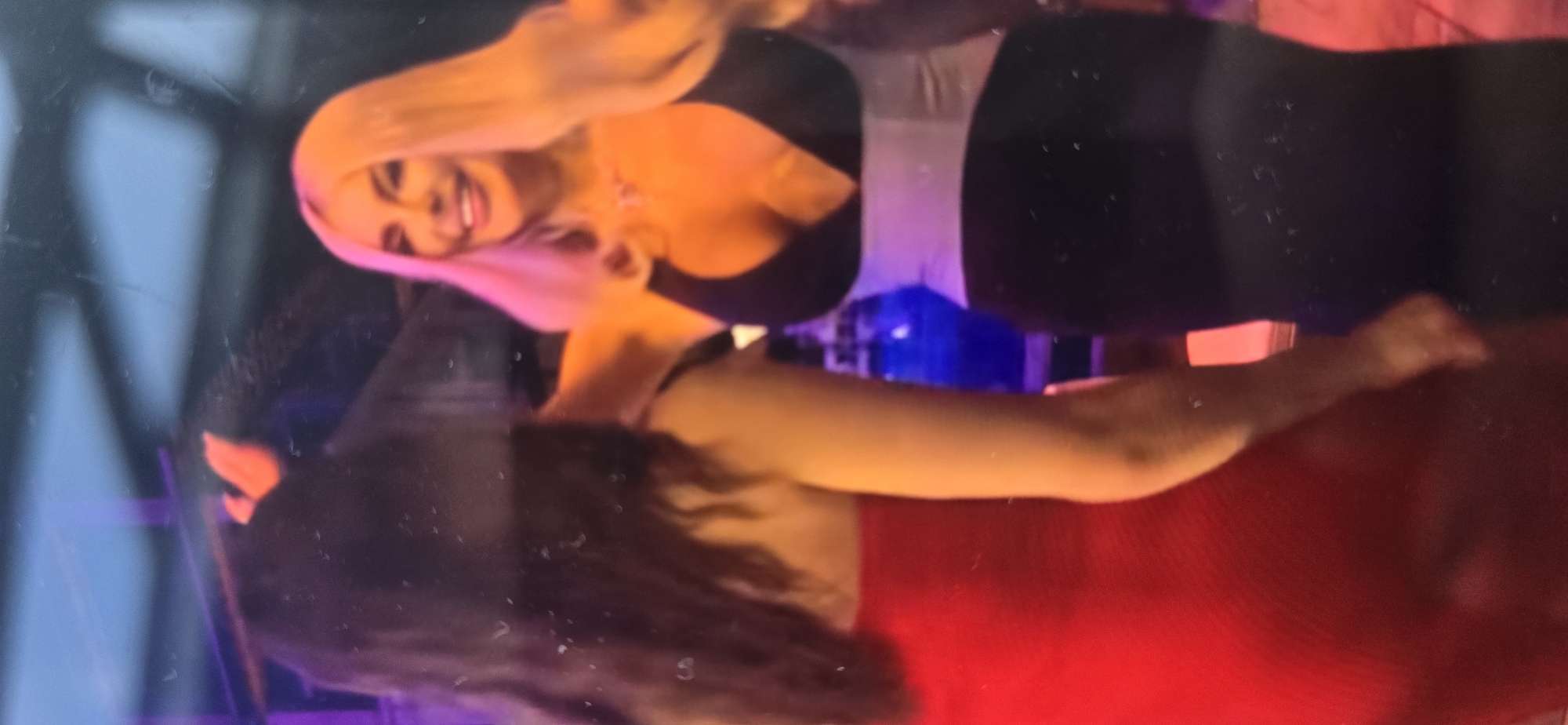 The image depicts two individuals, a woman with long dark hair wearing a red dress and another woman with blonde hair wearing a black dress with a silver sash. The woman in the black dress is smiling and has her arm around the woman in the red dress. They appear to be at an indoor event, possibly a party or celebration, with a dimly lit background that includes some colored lights. The image is grainy and appears to be a photograph taken from a screen. There is no information in the image to suggest the location is in the city of Kurgan, Russia.