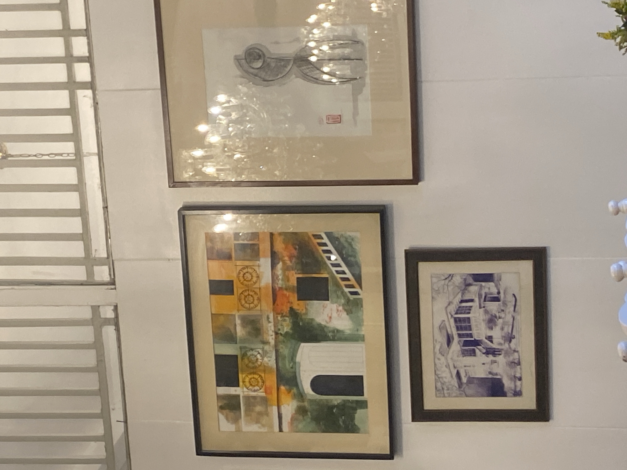 The image shows three framed artworks hanging on a white wall. The artwork on the left is a colorful painting depicting an architectural scene with a white arched doorway and a staircase. The artwork in the center right is a sketch of an abstract figure with a red stamp or seal. The artwork on the bottom left is a monochromatic sketch of a house. A chandelier is visible reflecting in the artwork on the right. The scene appears to be an indoor space, possibly a restaurant or a gallery, given the decor. There is no visible indication that the location is in Davao City, Philippines.