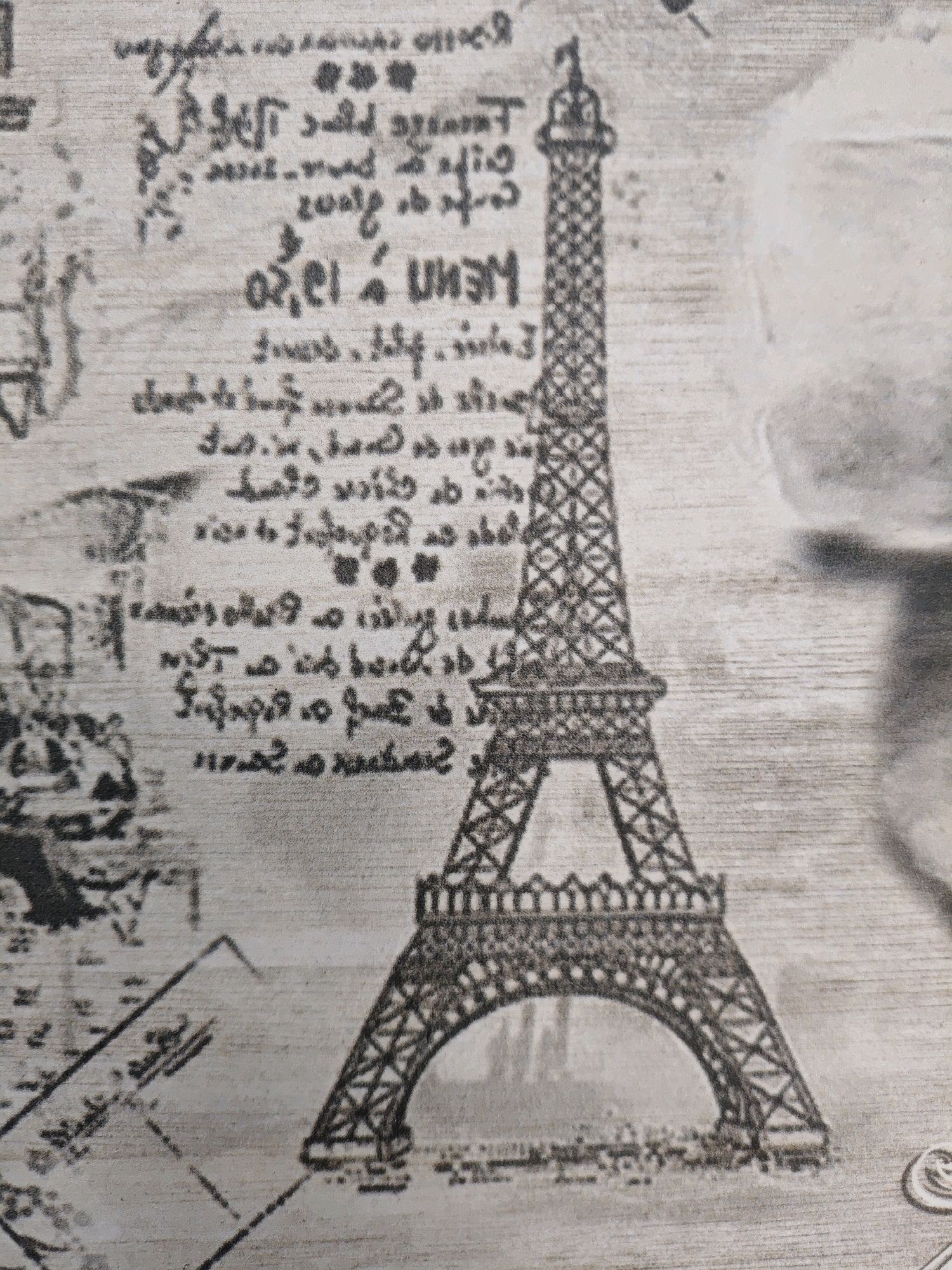 The image shows a close-up of a decorative surface, likely wallpaper or fabric, featuring a grayscale print of the Eiffel Tower and script. The script is handwritten and appears to be in French, with some words visible such as "Paris" and possibly "Eiffel". The print is stylized and aged, giving it a vintage feel. The background has a wood-grain texture. There are no people or discernable actions taking place. The image does not depict any location in the city of Ninotsminda, Georgia.