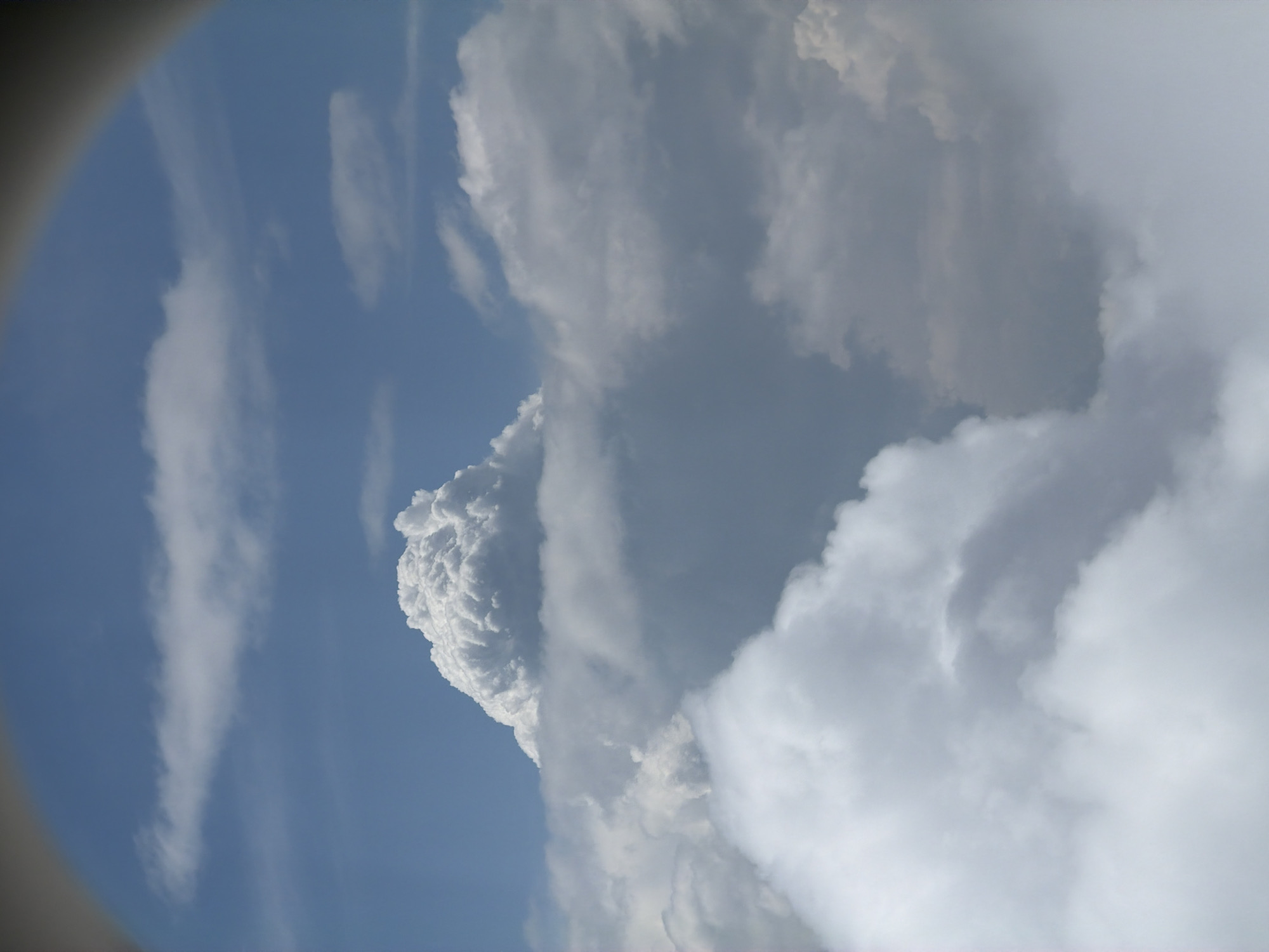 The image displays a view of clouds from an aircraft window. The clouds are dense and white, with some areas appearing darker and more shadowed, suggesting varying altitudes and densities. The sky is a clear blue. The scene appears to be an aerial view, looking down on a cloudscape. There are no discernible buildings, landmarks, or geographical features that would allow for identification of a specific location like Khimki, Russia. The image is devoid of people, objects, or actions.