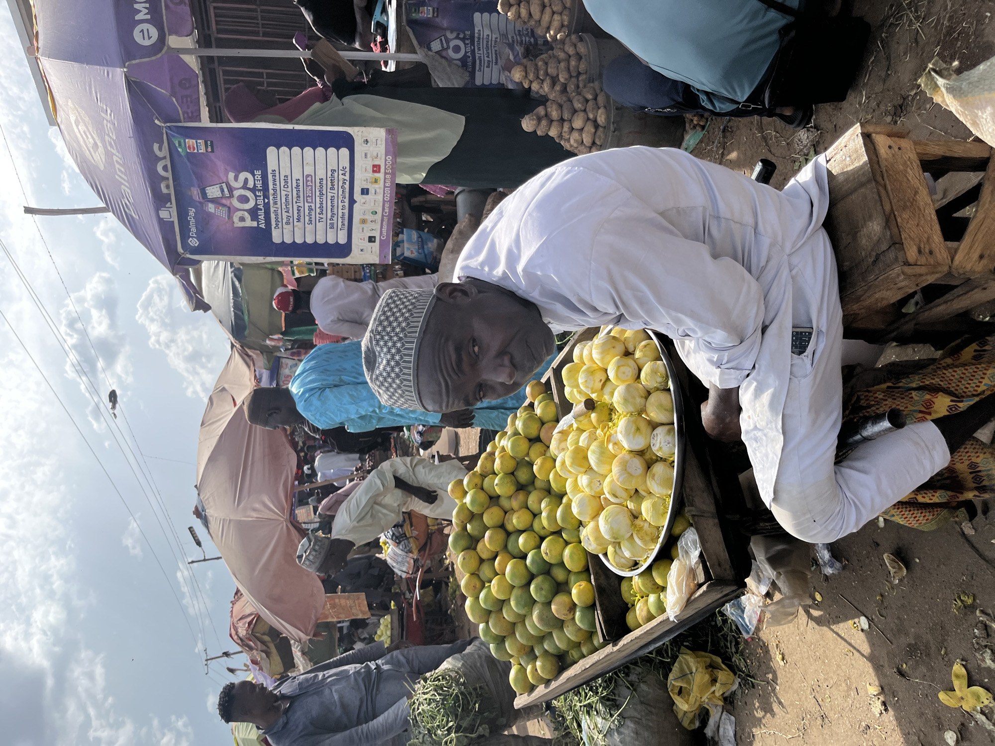 The image captures a busy outdoor market scene, likely in Rafin Par, Nigeria, under a bright, cloudy sky. In the foreground, a man wearing a white traditional outfit and a patterned skullcap sits on a wooden stool. He is positioned next to a large cart overflowing with green and yellow fruits, possibly oranges or tangerines, and a metal basin filled with peeled yellow fruits, possibly lemons. To the right of the man, several stacks of potatoes are visible in buckets. Behind the man and the fruit cart, other market-goers and stalls are present, with various umbrellas providing shade. A prominent purple umbrella with the "PalmPay POS" logo is visible, displaying information about available services such as deposits, withdrawals, money transfers, and bill payments. The ground is dusty and littered with debris. Other individuals in traditional attire are seen amidst the stalls and in the background, contributing to the bustling atmosphere of the market.