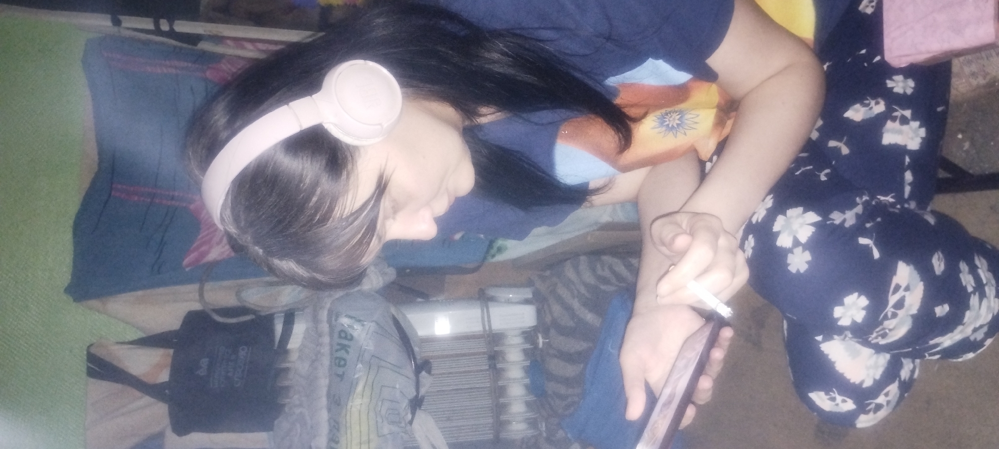 A woman with dark hair, wearing pink headphones, is sitting and holding a smartphone and a cigarette. She is wearing a dark blue shirt and dark blue pants with a white floral pattern. The background appears to be indoors, with some laundry and bags visible. The scene is dimly lit.