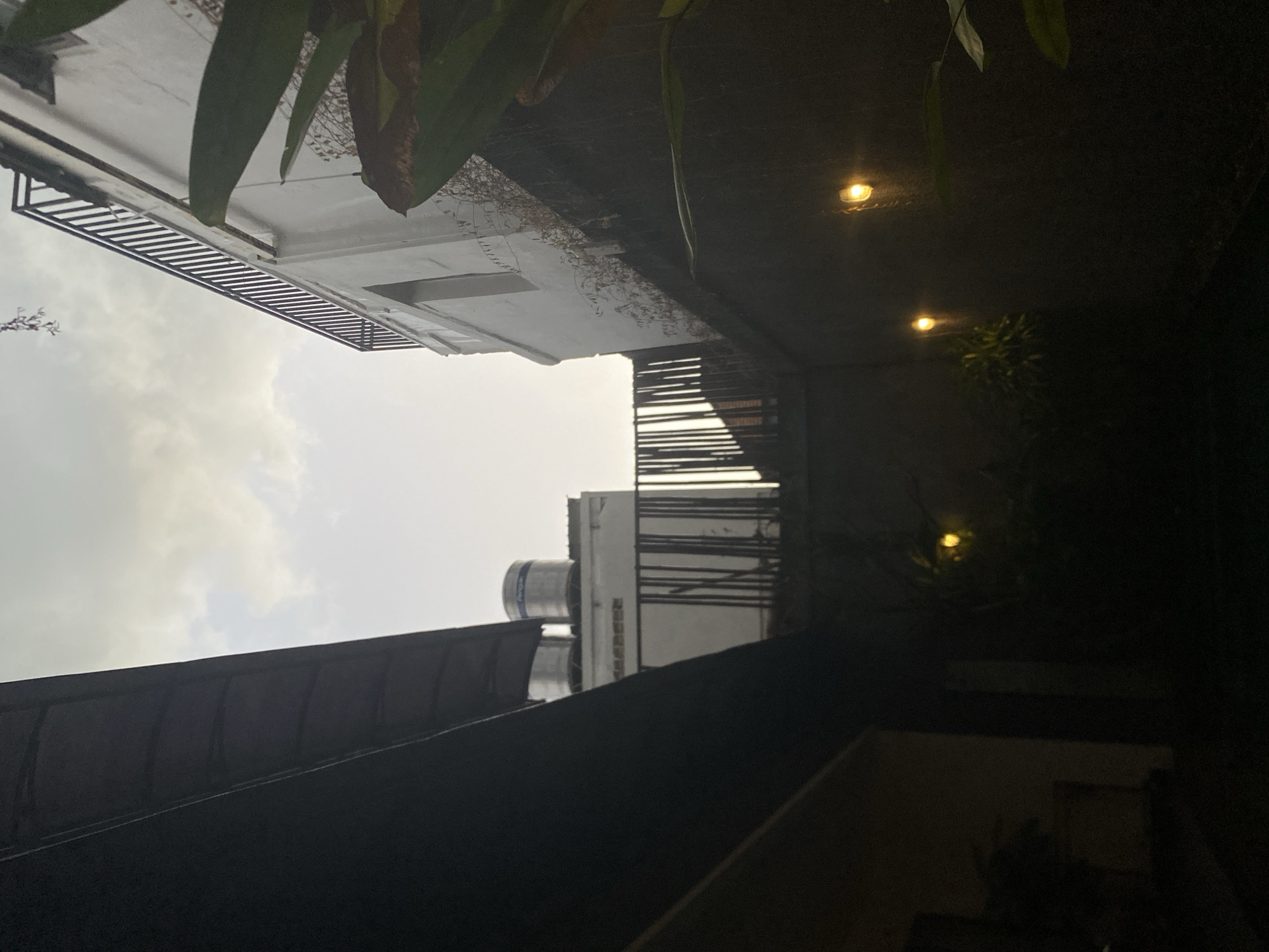 The image shows an outdoor courtyard at dusk or during a storm. In the foreground, a dark, sloped structure, possibly a roof or awning, occupies the left side of the frame. Beyond this, the scene opens up to reveal the sky, which is overcast with hints of light.
To the right of the dark structure, a white building with a black railing on its upper level is visible. Adjacent to this building, a fence or wall made of vertical wooden slats runs across the mid-ground. Two water tanks, one labeled "Penguin," are situated on top of another building behind this fence.
The lower part of the image is predominantly dark, suggesting it is at ground level or below. Two small, round lights with a warm yellow glow are visible on a dark wall on the right side of the frame. Some foliage, including large green leaves, is also present on the right. The overall impression is of a somewhat enclosed outdoor space, possibly a backyard or a private courtyard, with artificial lighting. There are no people visible in the image, nor any indication of specific activities. The presence of overcast skies and what appear to be streaks of rain on the wall suggest potentially inclement weather.