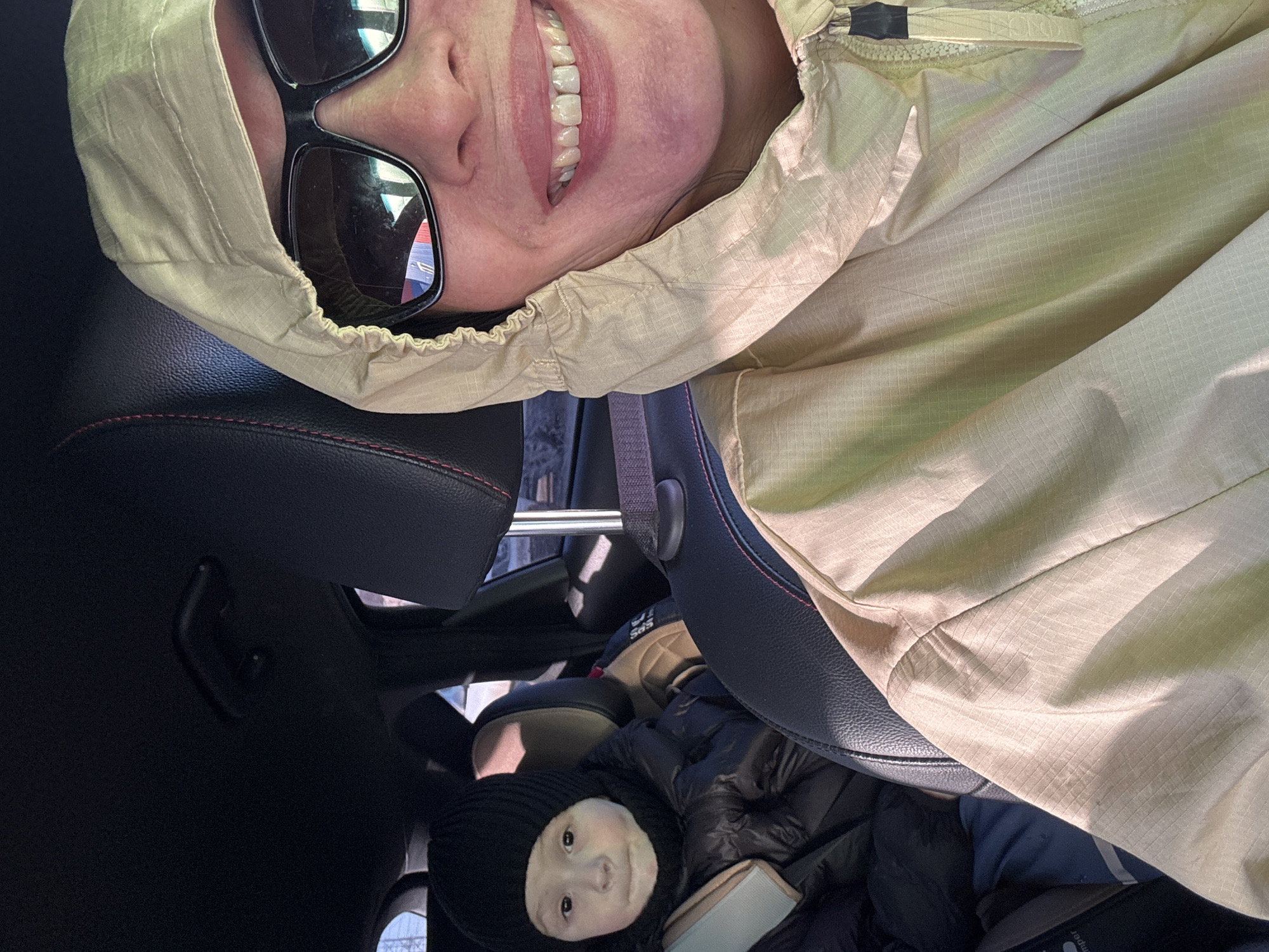A person wearing sunglasses and a beige jacket with a hood is smiling at the camera. The person is likely in a car, as suggested by the car seat and headrest visible in the frame. A child wearing a black knit cap is in a car seat below the person. The location is in Chaikovsky, Russia.