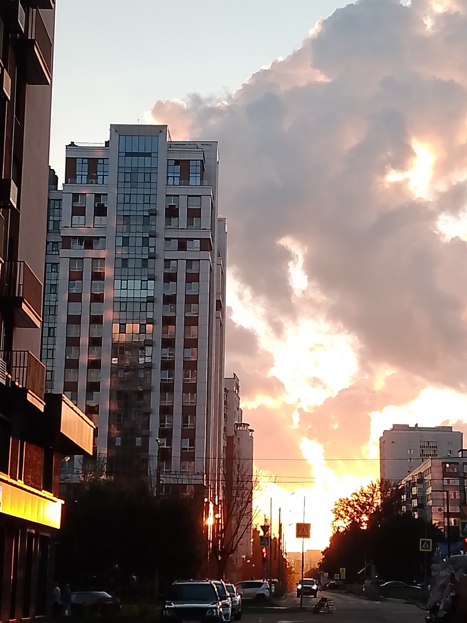 The image captures a street scene in Penza, Russia, during sunset. The sky is filled with dramatic clouds illuminated by the setting sun, casting a warm, orange glow. In the foreground, a modern, multi-story building with a facade of white panels and numerous windows dominates the left side of the frame. To its right, another tall building with a similar design is visible, reflecting the sunset's colors.
The street below is lined with trees and streetlights. Several cars are parked or driving on the road. Pedestrians are sparsely visible, and traffic signs, including pedestrian crossing signs, are present. The overall atmosphere is that of an urban environment transitioning from day to night, with the sunset creating a striking visual backdrop. The architectural style suggests a relatively modern development in the city.