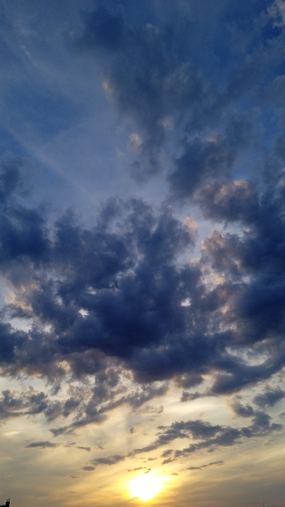 A vertical, low-angle, medium shot captures the expanse of a sky at sunset, with a bright, glowing sun radiating from the lower right quadrant. The sun, a brilliant orb of white and yellow light, is partially obscured by a wispy layer of clouds. Above the sun, the sky transitions through a spectrum of colors, from warm oranges and yellows near the horizon to a pale, hazy blue higher up. This area is populated by scattered, thin clouds that are illuminated by the setting sun, creating a soft, ethereal glow.
The middle and upper portions of the image are dominated by a dramatic display of dark, turbulent clouds. These clouds are a deep, bruised blue, tinged with subtle hints of pink and grey where they catch the fading light. They are densely packed, creating a sense of depth and texture against the lighter sky. Some of these clouds are thick and heavy, suggesting the possibility of rain, while others are thinner and more fragmented, allowing glimpses of the blue sky peeking through. The clouds in the upper section appear to be moving, with streaks of lighter cloud visible, possibly indicating wind patterns at higher altitudes.
Along the very bottom edge of the frame, a dark, silhouetted structure is visible on the left. It is a narrow, vertical object, possibly a building or a tree, appearing as a stark contrast to the luminous sky above. The overall impression is one of a dynamic and awe-inspiring sunset, characterized by dramatic cloud formations and the intense glow of the setting sun. The location is described as Unknown, Unknown.
