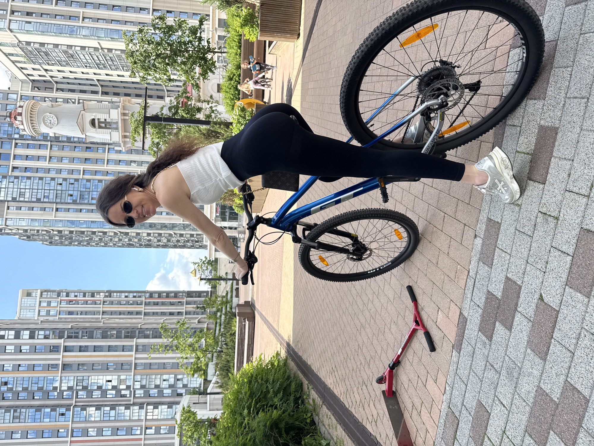 A young woman is captured posing playfully with a blue mountain bike in an urban outdoor setting in Moscow, Russia. She is leaning forward over the bike's handlebars in a stretched position, looking back at the camera with a wide smile. She has long, dark, wavy hair, and is wearing a white ribbed sleeveless top, form-fitting black leggings, and dark sunglasses. Gold earphones are visible in her ears, and a gold chain necklace is around her neck. A small brown handbag with a distinctive monogram pattern resembling Louis Vuitton is draped over the bike's seat post. Her right foot, clad in a chunky silver and white sneaker with a cross-like design, rests on the ground. The bike, which has "GT" visible on its frame and "WTB" on the tire, features orange reflectors on its spokes and silver gears, possibly Shimano.
The scene is brightly lit by natural daylight, indicating clear and sunny weather. The ground is paved with light brown and grey interlocking pavers. In the mid-ground, a red kick scooter lies on the ground near the bike's front wheel. Further back, two other women are visible near benches, suggesting a public area or park within a residential complex. The background is dominated by tall, modern apartment buildings with numerous windows, reflecting the sky. A notable architectural feature is a clock tower building with a white facade and a red-domed top, positioned behind the woman and the bike. The overall atmosphere appears casual and joyful.