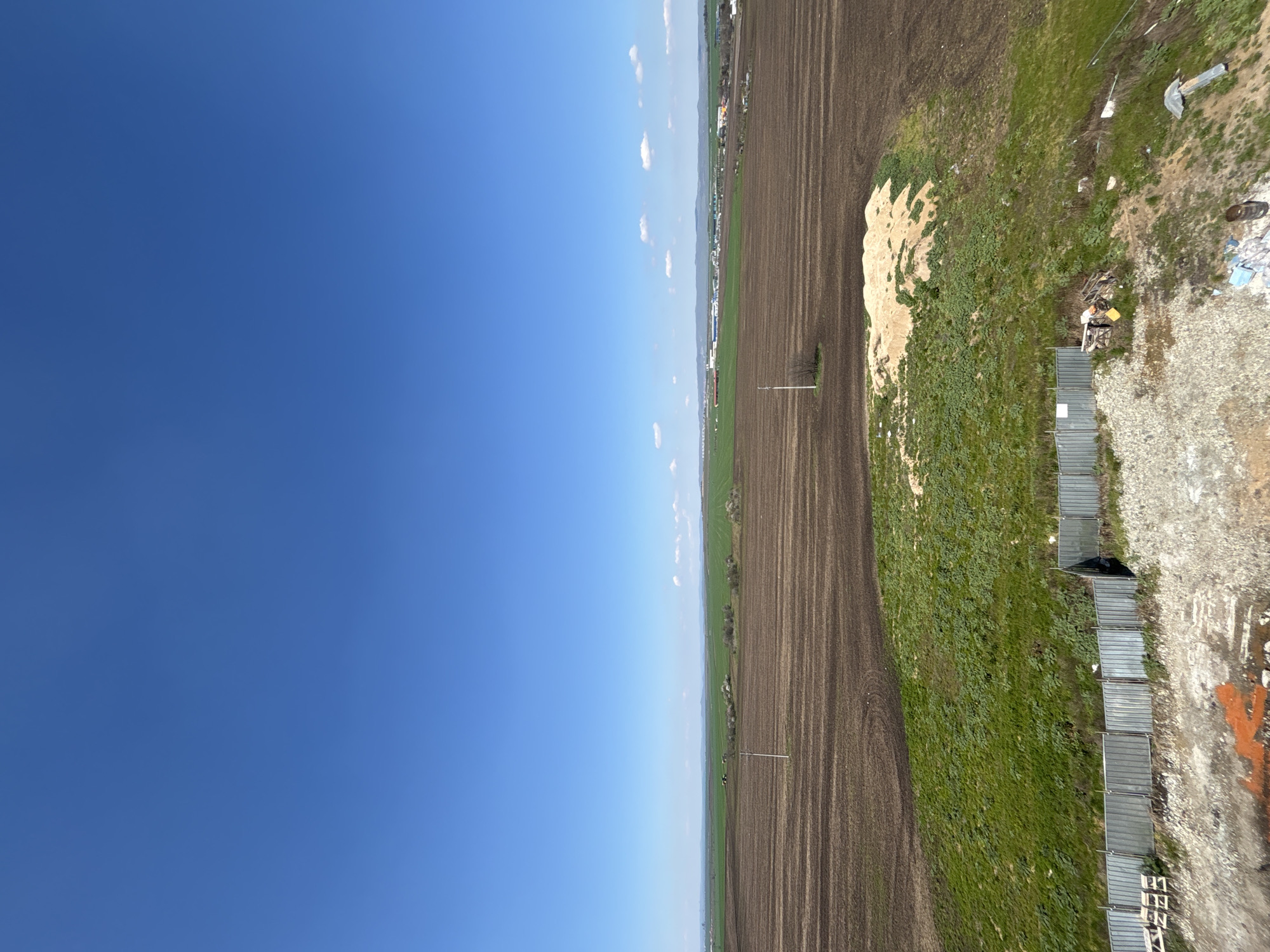 The image shows an aerial perspective of a landscape likely situated near Burgas, Bulgaria. The scene is divided between a large expanse of clear, blue sky and a terrestrial view of agricultural land. The land features freshly plowed fields with dark, furrowed soil alongside patches of green vegetation and grassy areas.
Along the right edge of the frame, there's evidence of human activity with elements like a metal fence, gravel, and what appears to be discarded materials or trash. There is also a light-colored pile of what might be sand or another construction material. The image includes details of a rural or peri-urban environment possibly on the outskirts of Burgas, reflecting a mix of agricultural and potentially industrial or construction-related activities. No people or identifiable human subjects are visible.